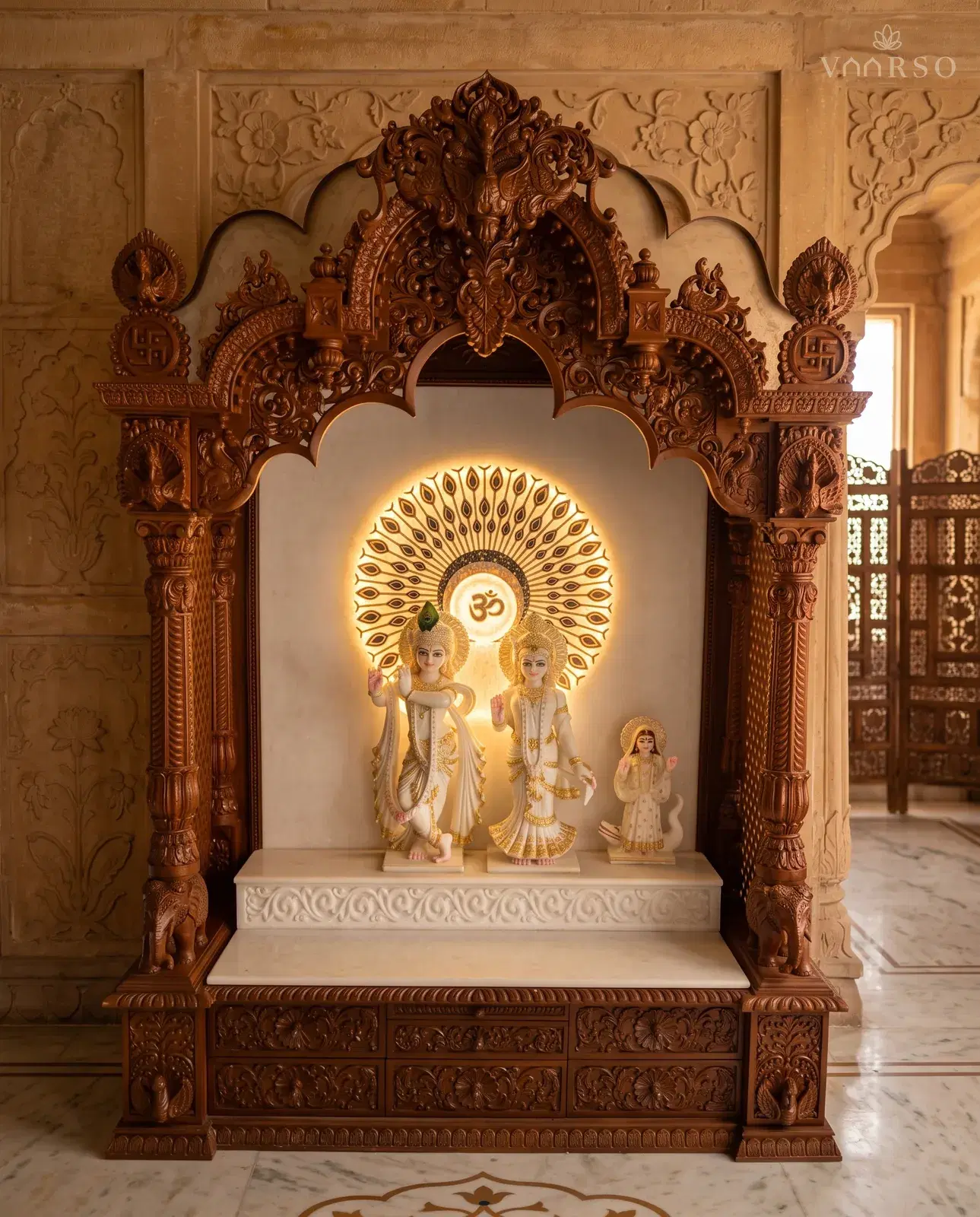 A mandir in a warm natural polish, a peacock-feather panel backlit behind three murtis, a carved arch on peacock columns, elephants at the base of the columns and carved drawers along the plinth