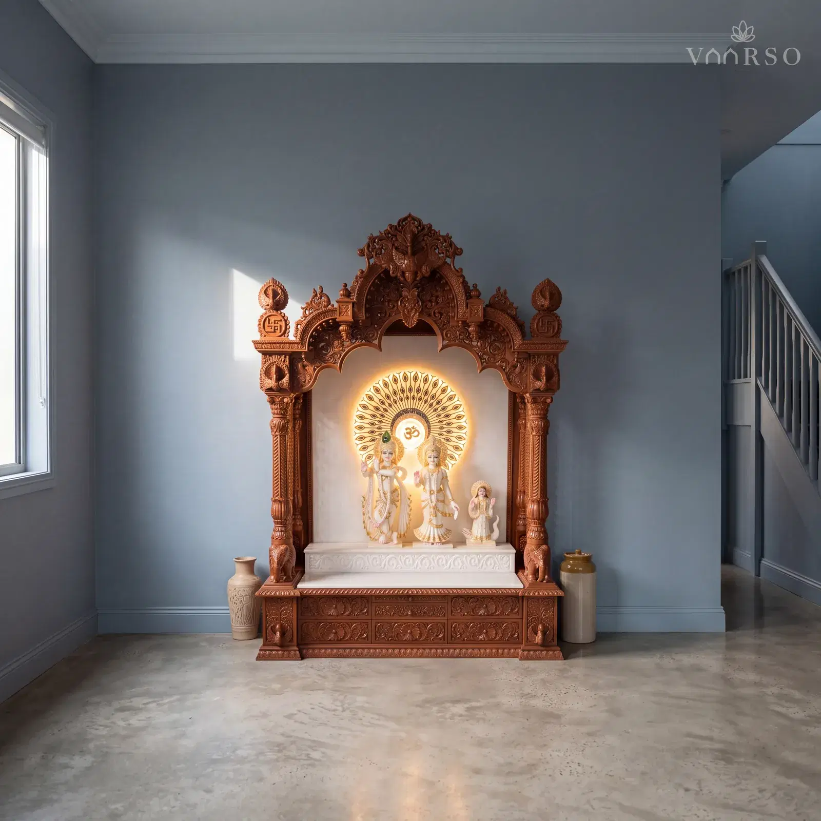 The same mandir in a narrow townhouse living room, the stair rising at the far end, polished concrete underfoot and a tall ceramic vessel on the floor to one side