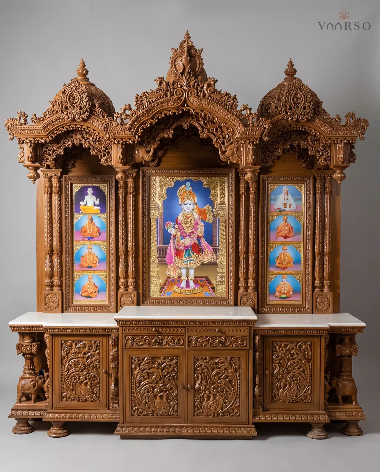 A wide mandir in a warm natural polish, two domes and a peacock crest over three bays, a large gilded picture at the centre with eight smaller framed figures either side, white marble shelves, and carved cupboards below