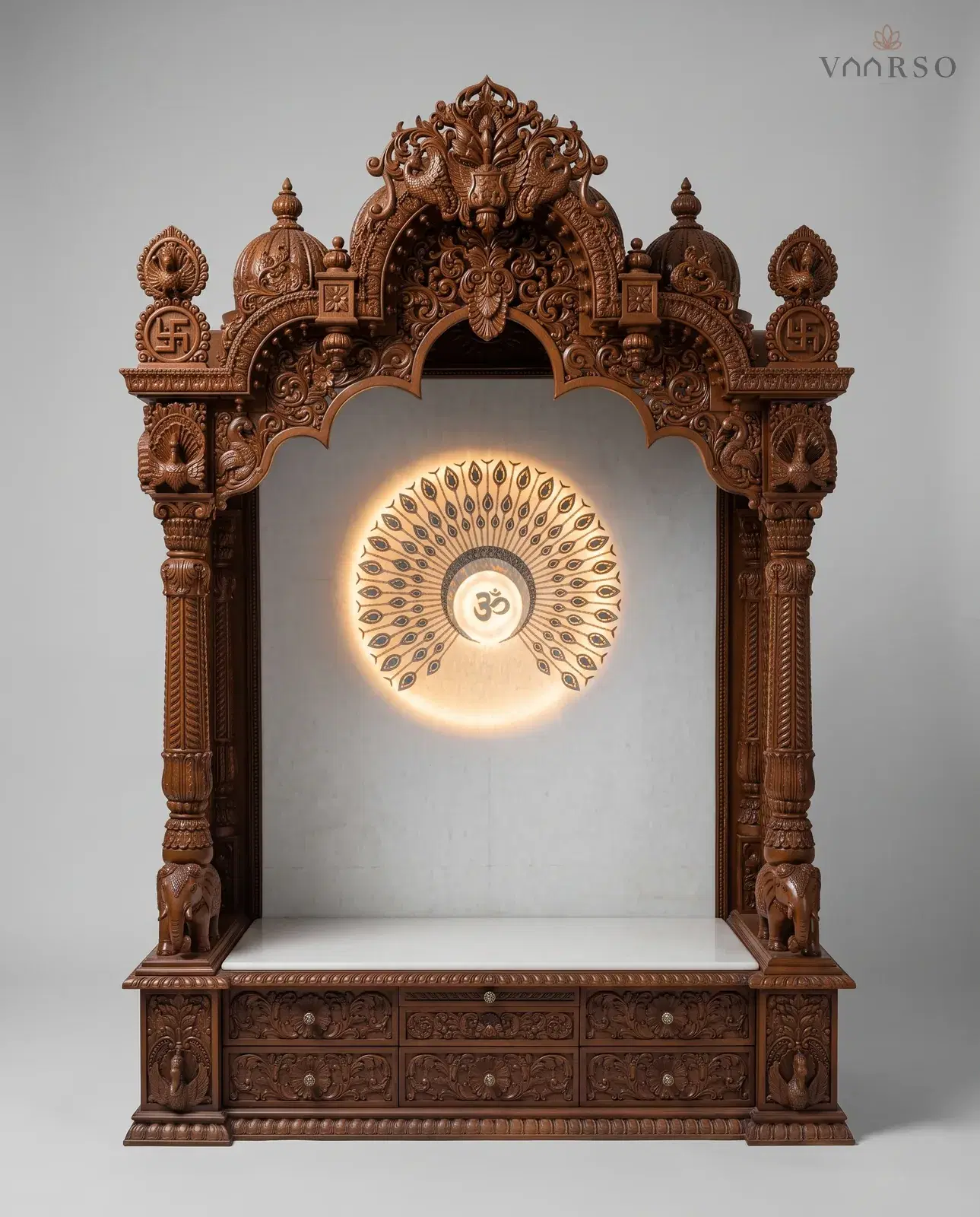 A tall mandir in a warm brown polish, a peacock crest and two domes over a scalloped arch, a backlit peacock-feather Om panel behind an empty white marble shelf, elephants at the columns, and six carved drawers across the plinth