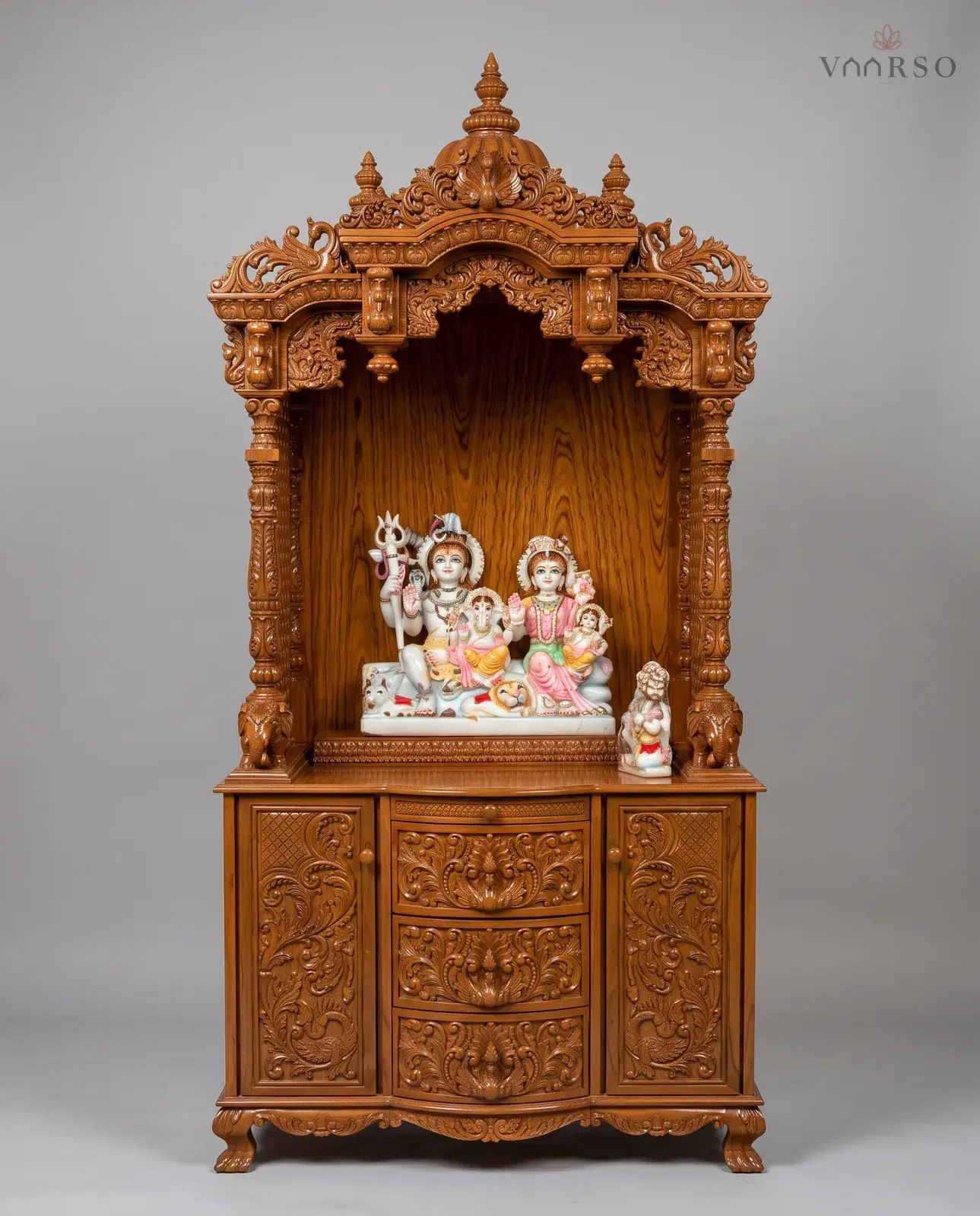 A tall mandir in a warm natural polish, a domed crest over a scrolled valance, a carved marble group standing in the open shrine, elephants at the foot of the columns, and curved drawers between two carved cupboards on claw feet