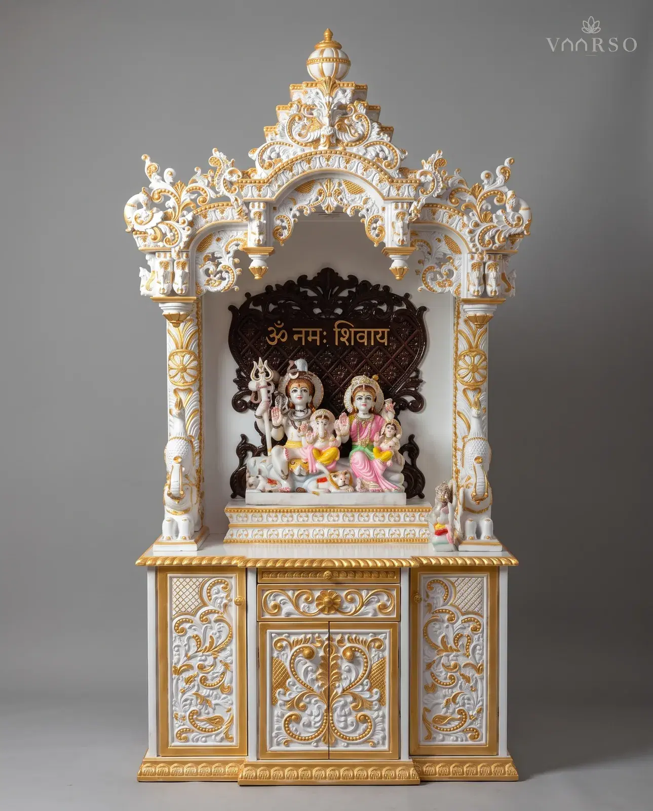 A mandir in white polish picked out in gold, a stepped crest over a pierced arch, a dark inscribed backpiece behind a seated marble group, white elephants at the columns, and a drawer over gilded cupboard doors