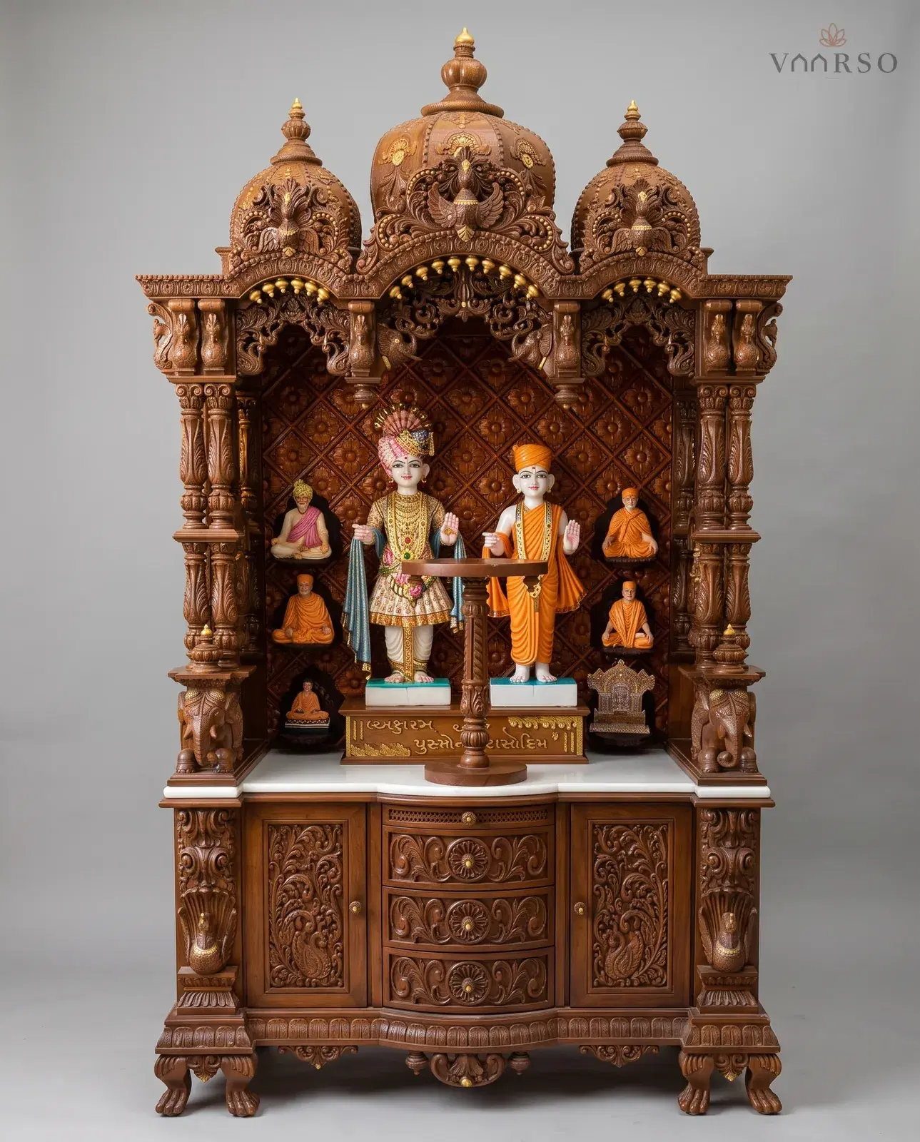 A tall mandir in a warm brown polish, three domes with gilded bud fringes over the arches, murtis standing before a diamond-patterned backboard with seated figures on the side shelves, elephants at the columns, and curved drawers between carved cupboards on claw feet