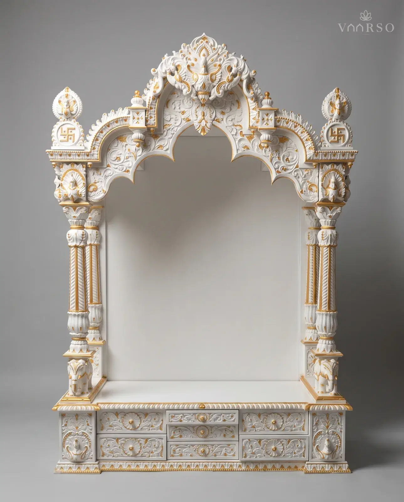 A mandir in white polish picked out in gold, a scrolled crest between two swastika roundels, a scalloped arch on twin turned columns with elephants at their feet, an empty shrine, and drawers across the carved plinth