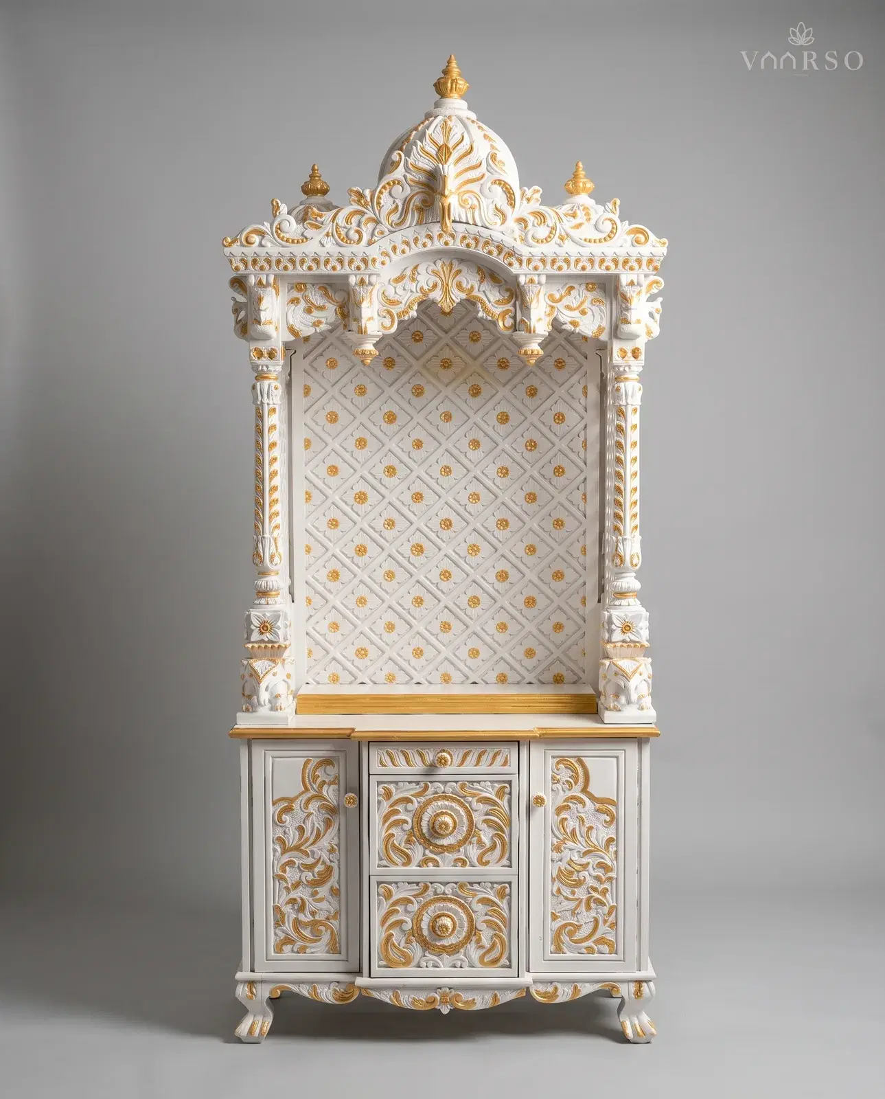 A tall mandir in white polish picked out in gold, a ribbed dome over a scrolled cornice, a diamond-patterned backboard studded with gold flowers behind the empty shrine, and a drawer over two carved cupboards on claw feet