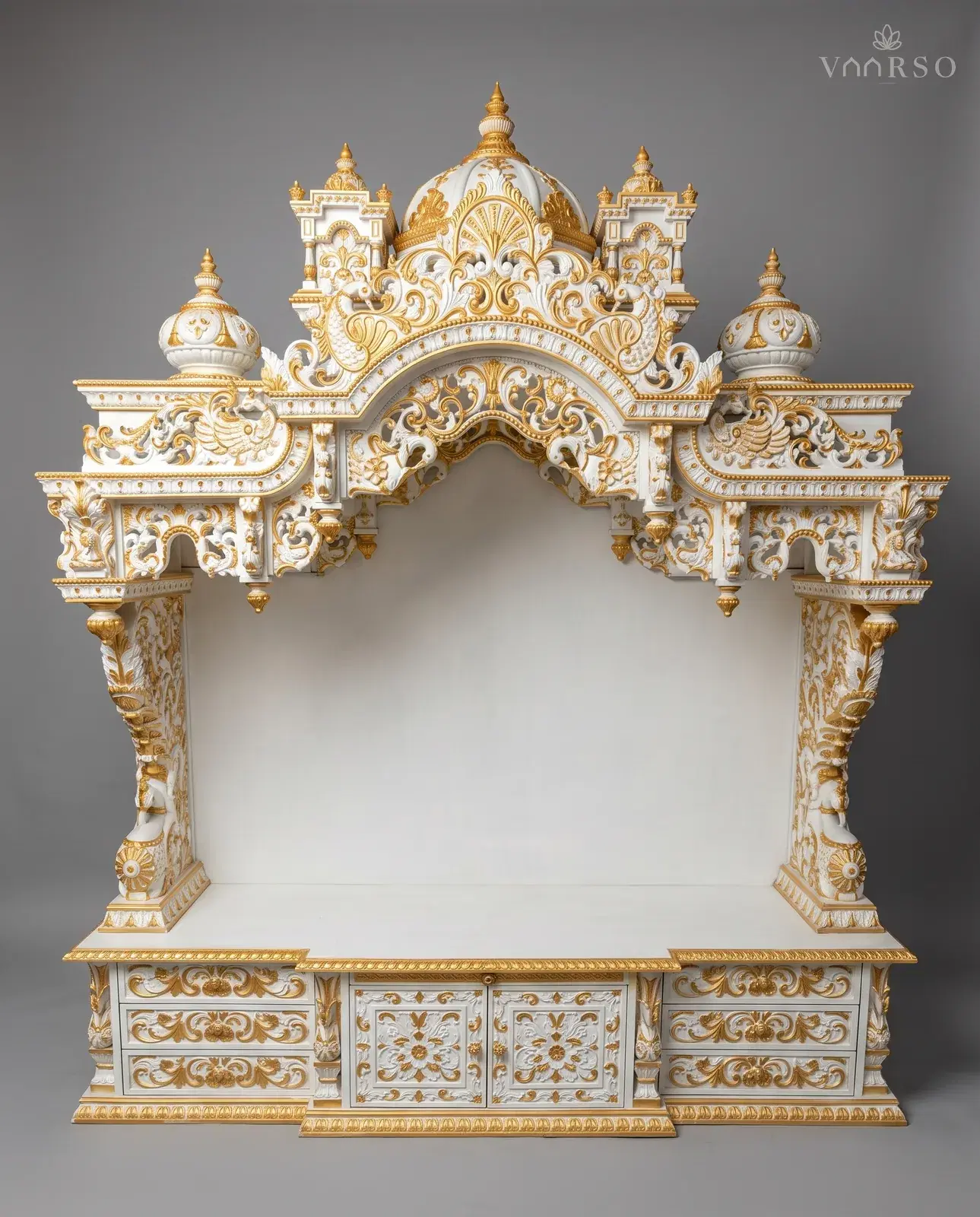 A wide mandir in white polish picked out in gold, a dome and two kiosks over a deeply pierced arch, peacocks in the side panels, an empty shrine, and drawers either side of two carved cupboard doors