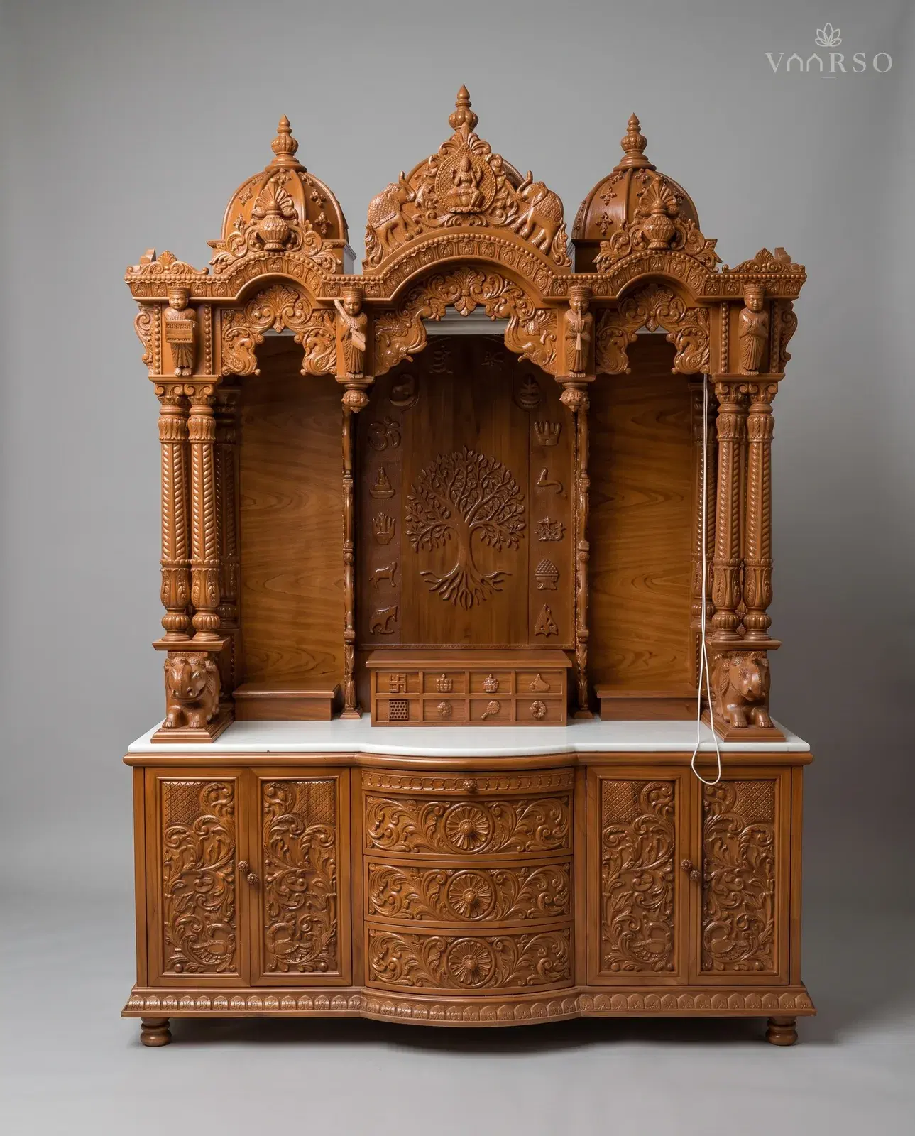A wide mandir in a warm natural polish, three domes over three bays with carved figures standing on the brackets, a tree of life carved into the back panel above a bank of small drawers, elephants on the marble shelf, and carved drawers between two cupboards