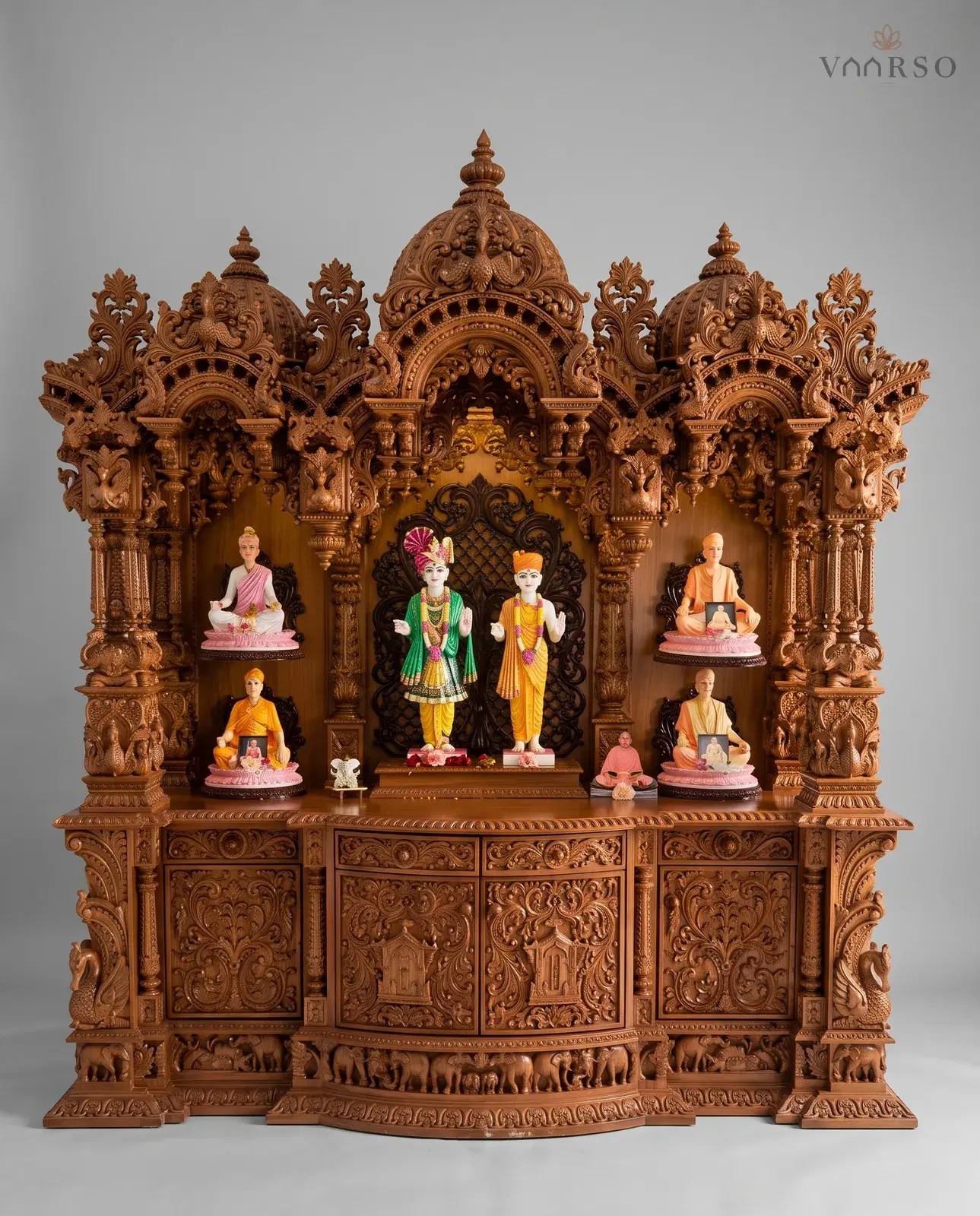 A wide mandir in a warm natural polish, three carved domes over a scrolled cornice, murtis standing in the central arch with seated figures on the side shelves, deeply carved cupboard doors below and a frieze of elephants around the plinth