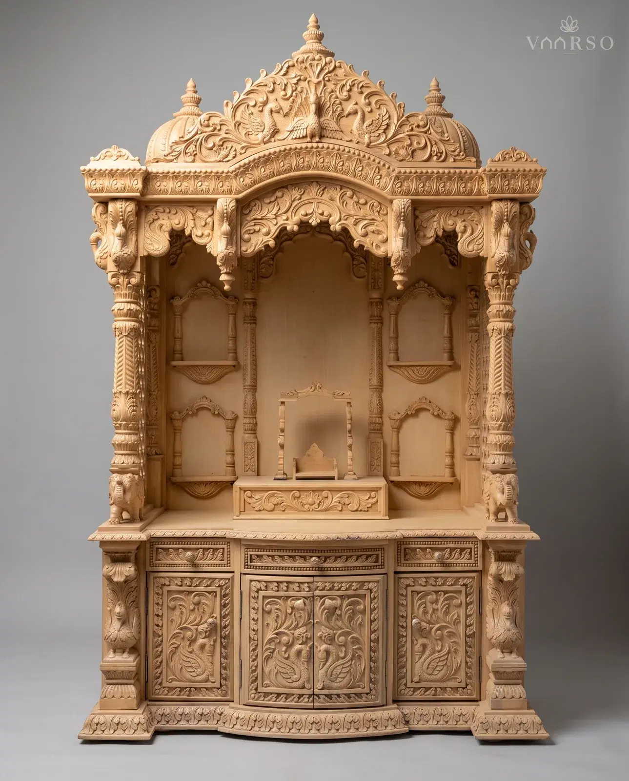 A tall mandir in bare unfinished timber, peacocks carved into the domed crest, four empty niche shelves either side of a small carved throne, and drawers over peacock-carved cupboard doors