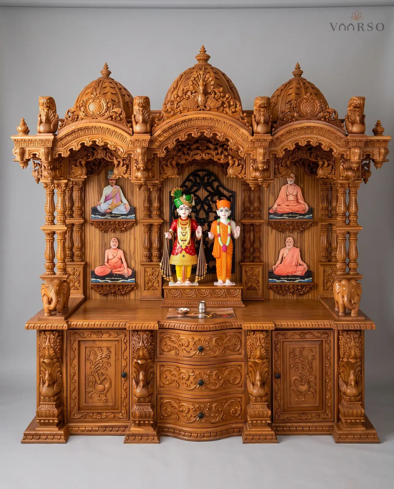 A wide mandir in a warm natural polish, three domes on lion brackets over a carved valance, framed portraits on the side panels, elephants at the outer corners, and serpentine drawers between two carved cupboards