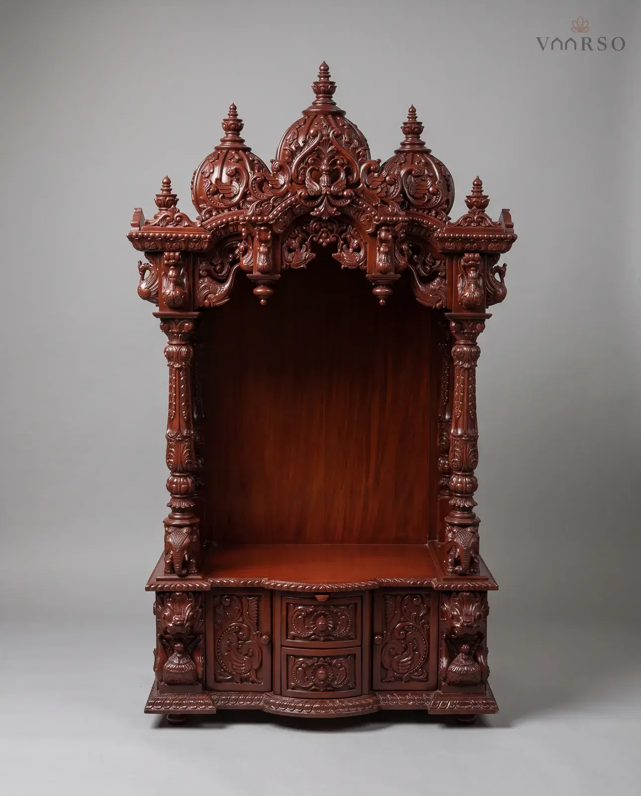 A tall mandir in a deep red-brown polish, three carved domes over a pierced arch, an empty shrine on turned columns with elephants at their feet, and a curved drawer between two carved cupboard doors
