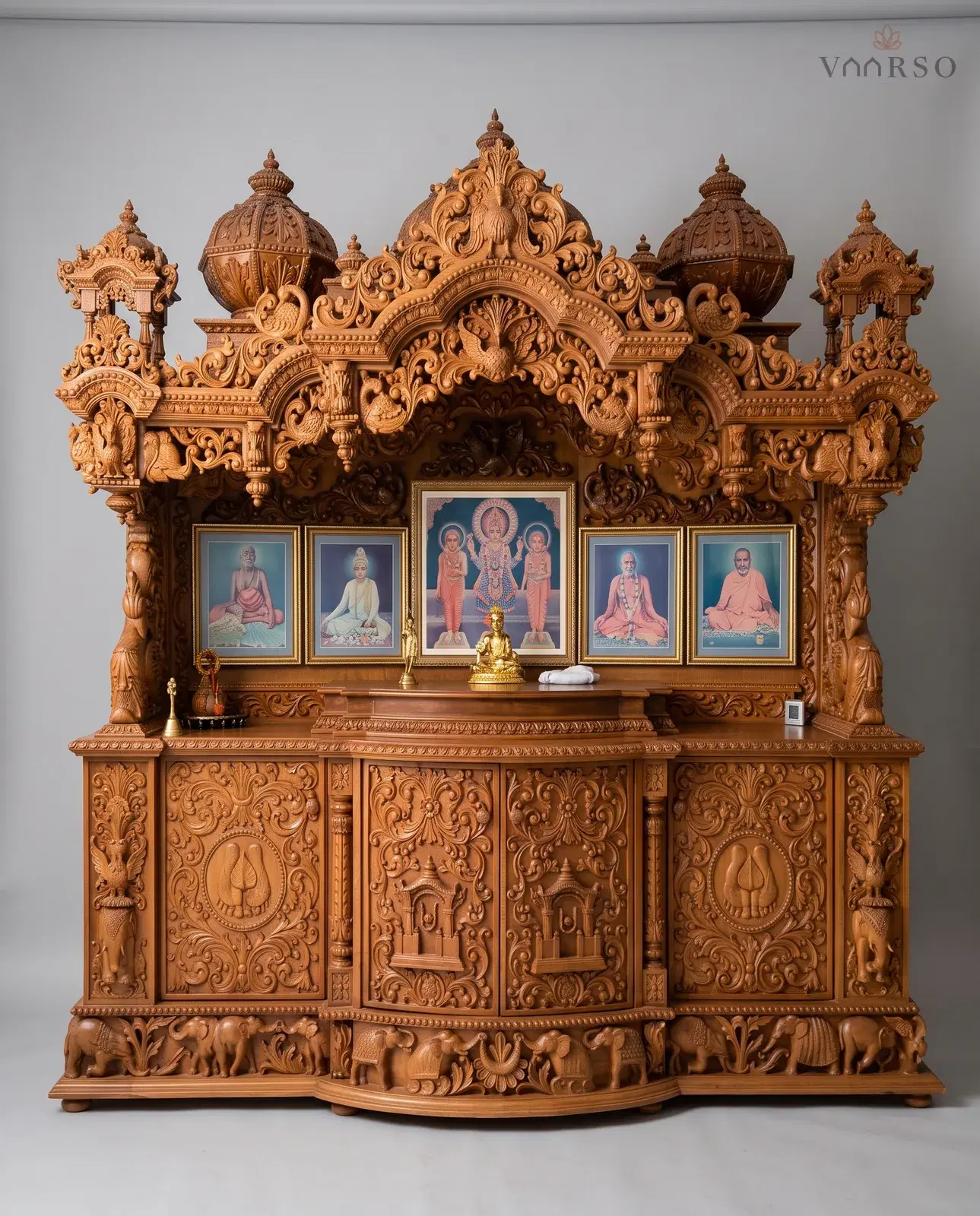 A wide mandir in a warm natural polish, domes and side kiosks above a deep carved cornice, five framed portraits standing along the shrine, and a run of carved cupboards over a plinth of elephants