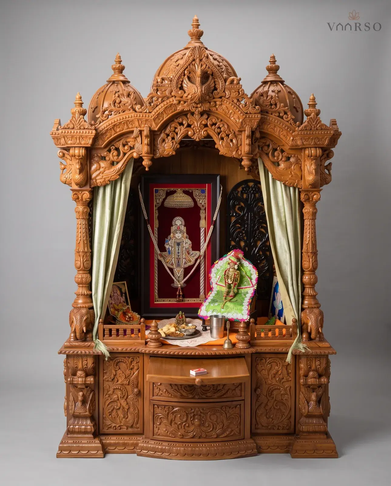 A mandir in a warm natural polish with pale green curtains drawn back at the columns, a framed picture and dressed murti inside, a low railing to the shelf, and a curved drawer over carved cupboard doors