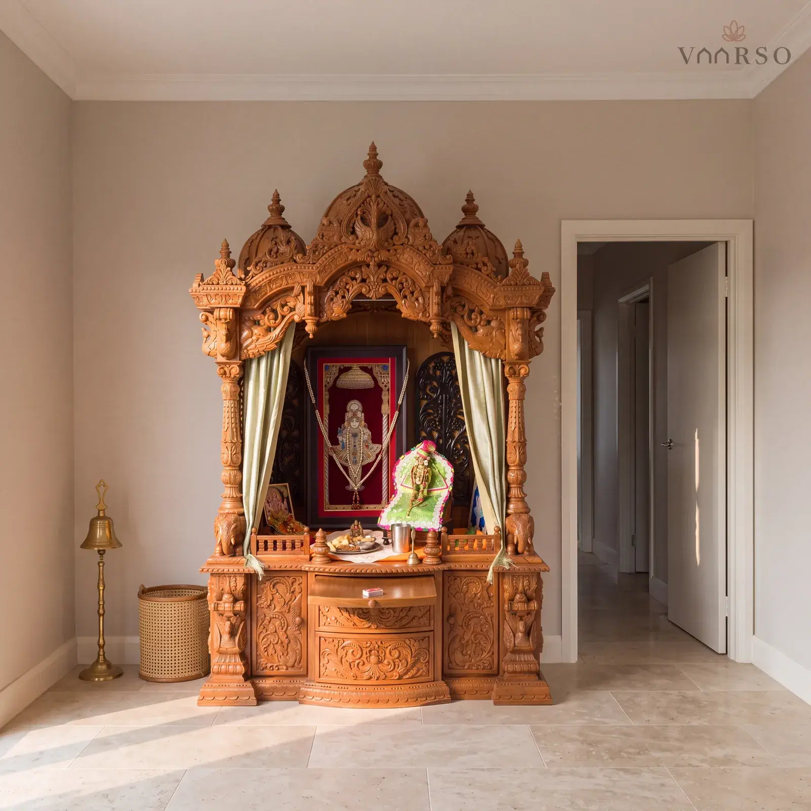 The same mandir in a small pooja room, standing in its alcove, honed travertine underfoot and a brass bell on a floor stand and a cane basket beside it