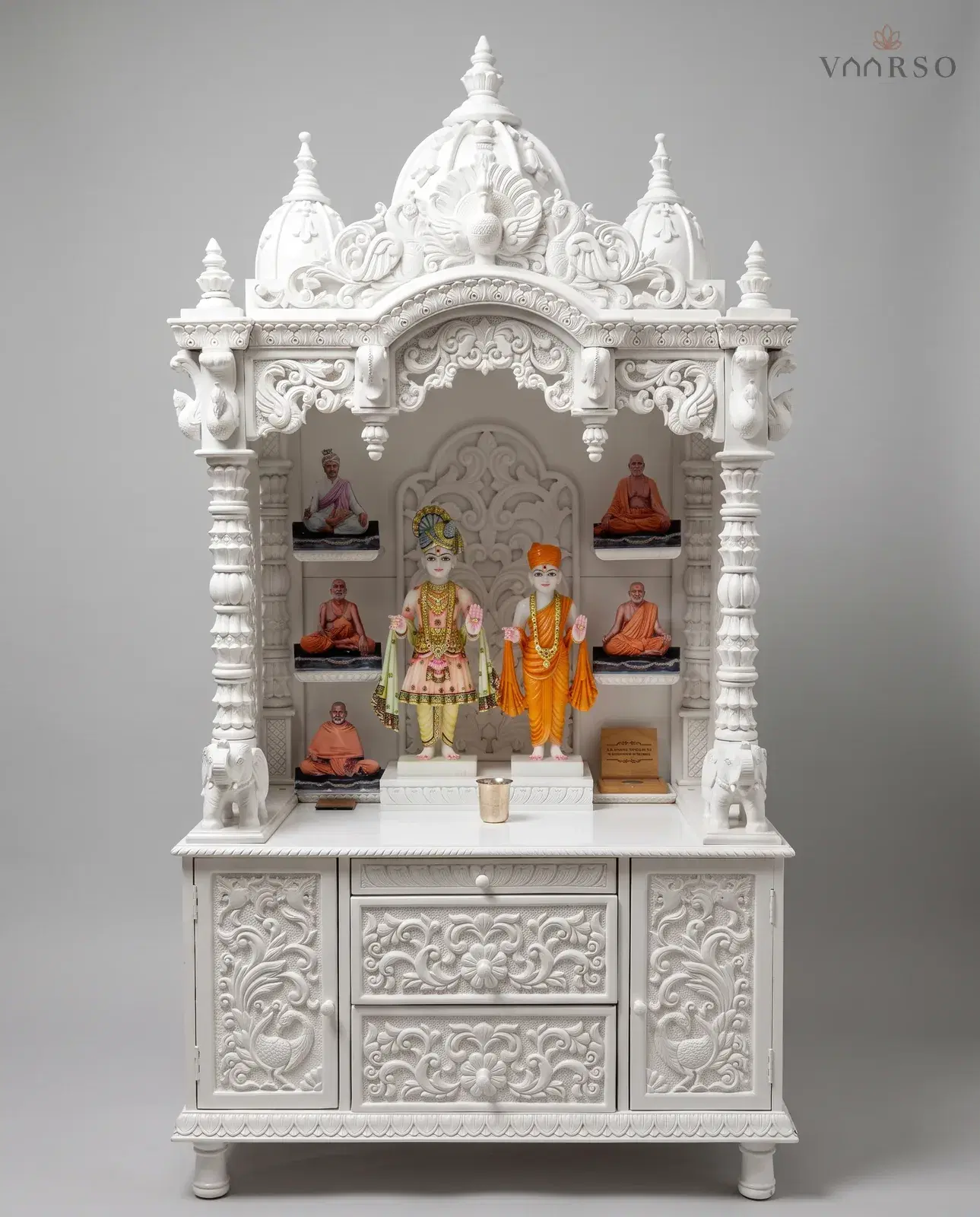 A mandir in a plain white polish, three domes over a carved valance, murtis standing before a pierced backpiece with framed portraits on the side shelves, white elephants at the columns, and carved drawers between two cupboards