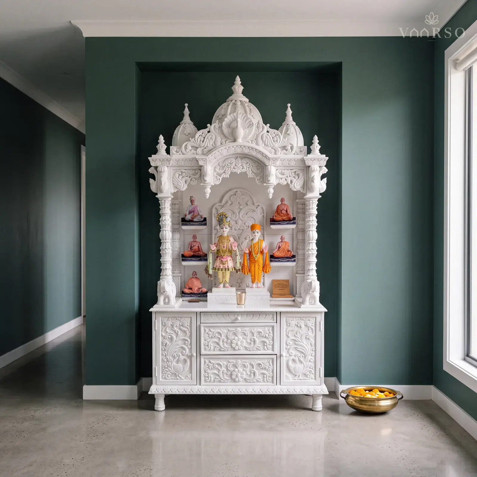 The same mandir in a wide hallway nook, set into the recess, polished concrete underfoot and a brass urli of floating marigolds on the floor
