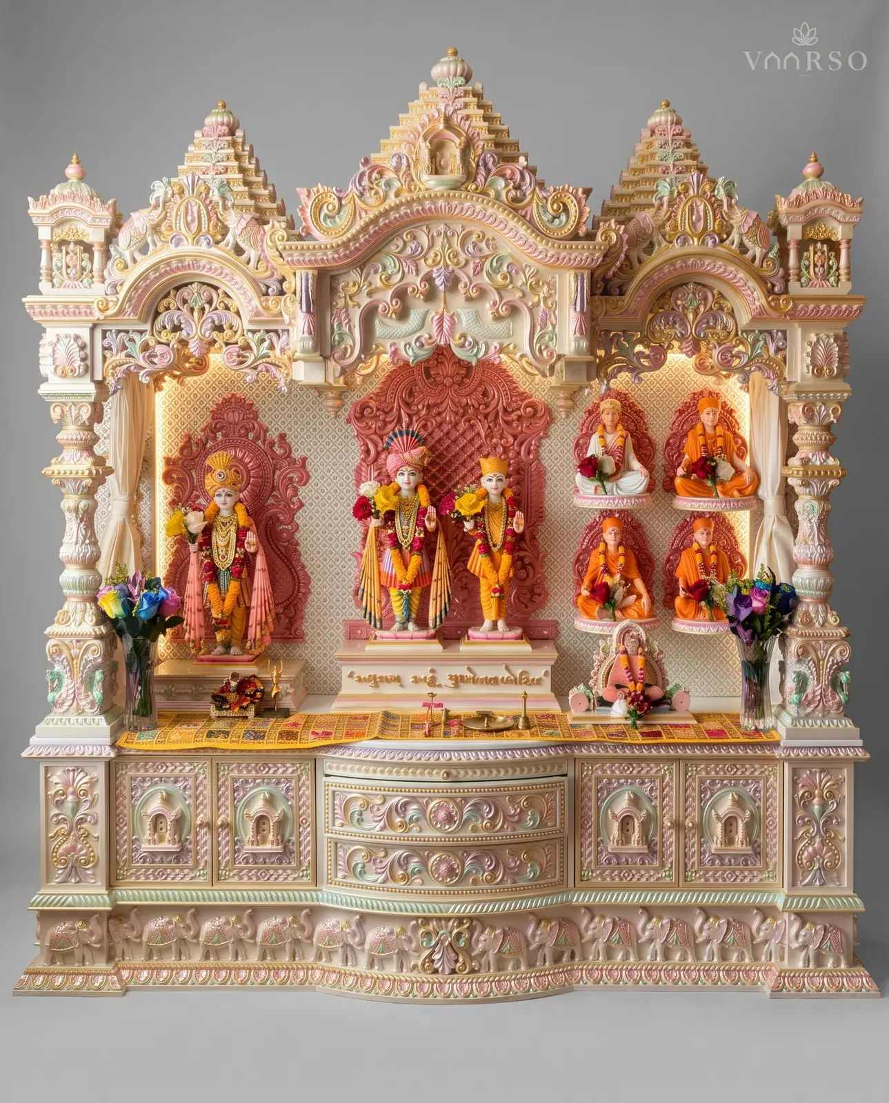 A wide mandir in cream polish picked out in pastel colours and gold, three stepped shikhars over a scrolled crest, three backlit bays holding murtis and seated figures, and carved drawers over a plinth of elephants