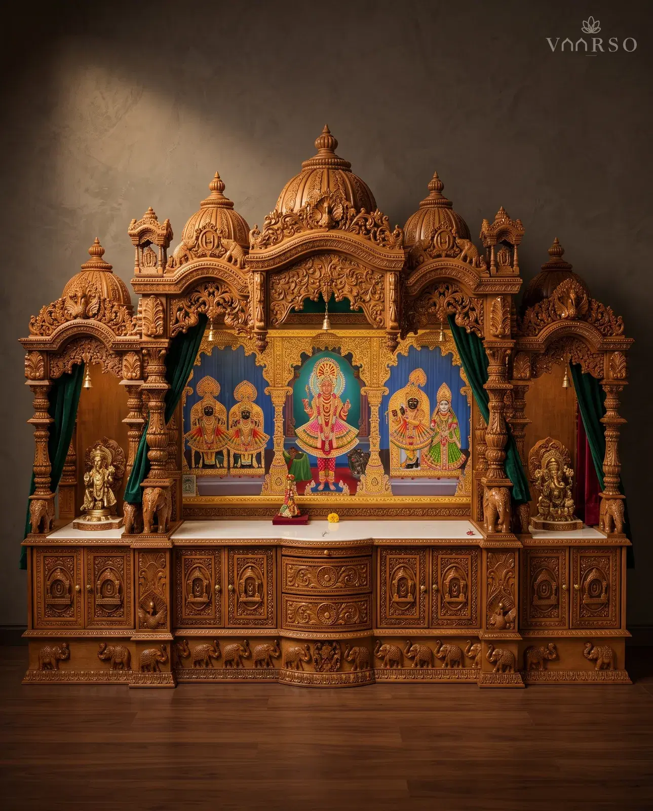A wide mandir in a warm natural polish, five domes and side kiosks across the crest, green curtains drawn back at the side bays, a painted shrine panel at the centre, brass figures on a white marble shelf, and carved cupboards over a band of elephants