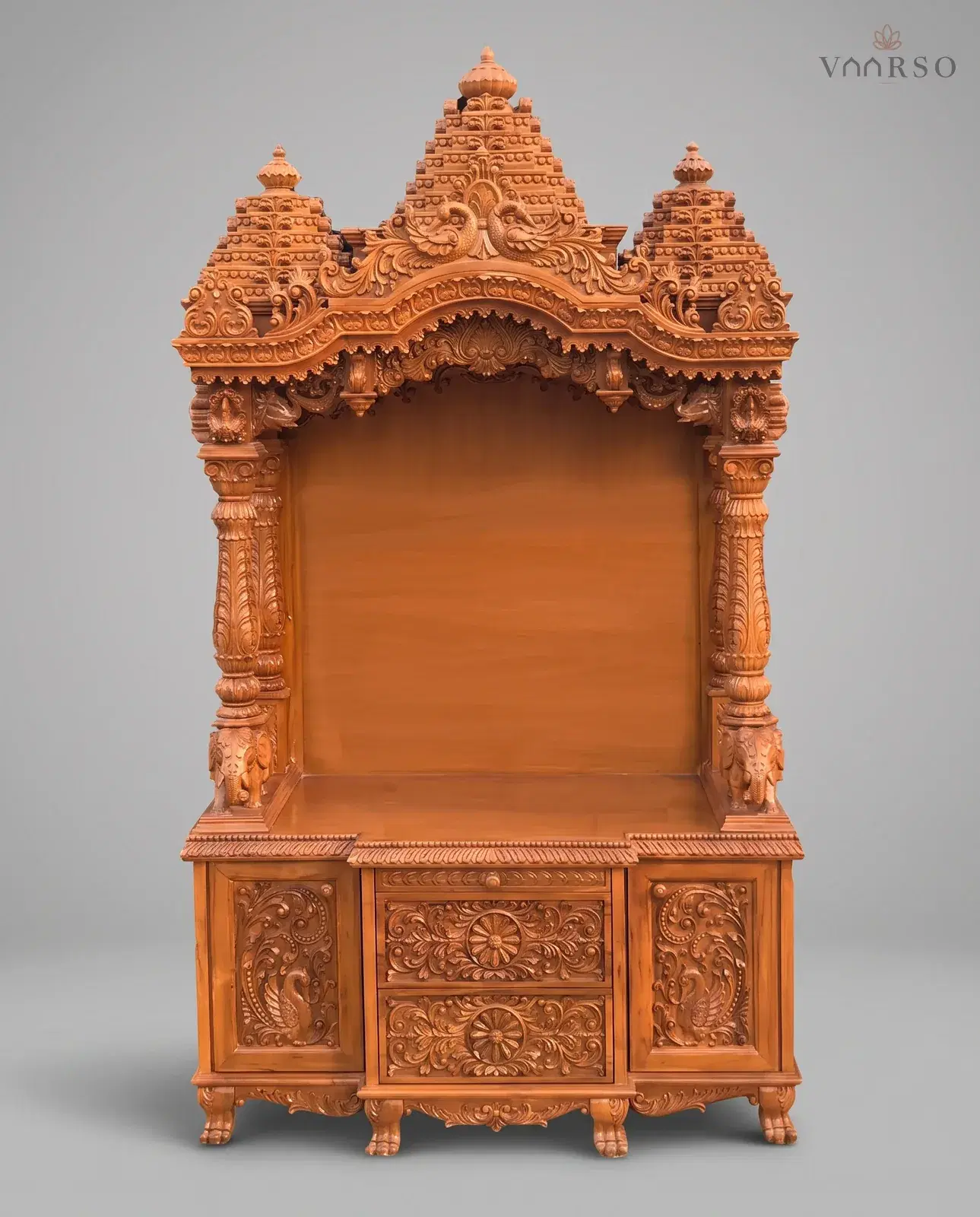 A tall mandir in a warm natural polish, three stepped shikhars with peacocks over a scalloped arch, an empty shrine on carved columns with elephants at their feet, and two drawers between peacock-carved cupboards on claw feet