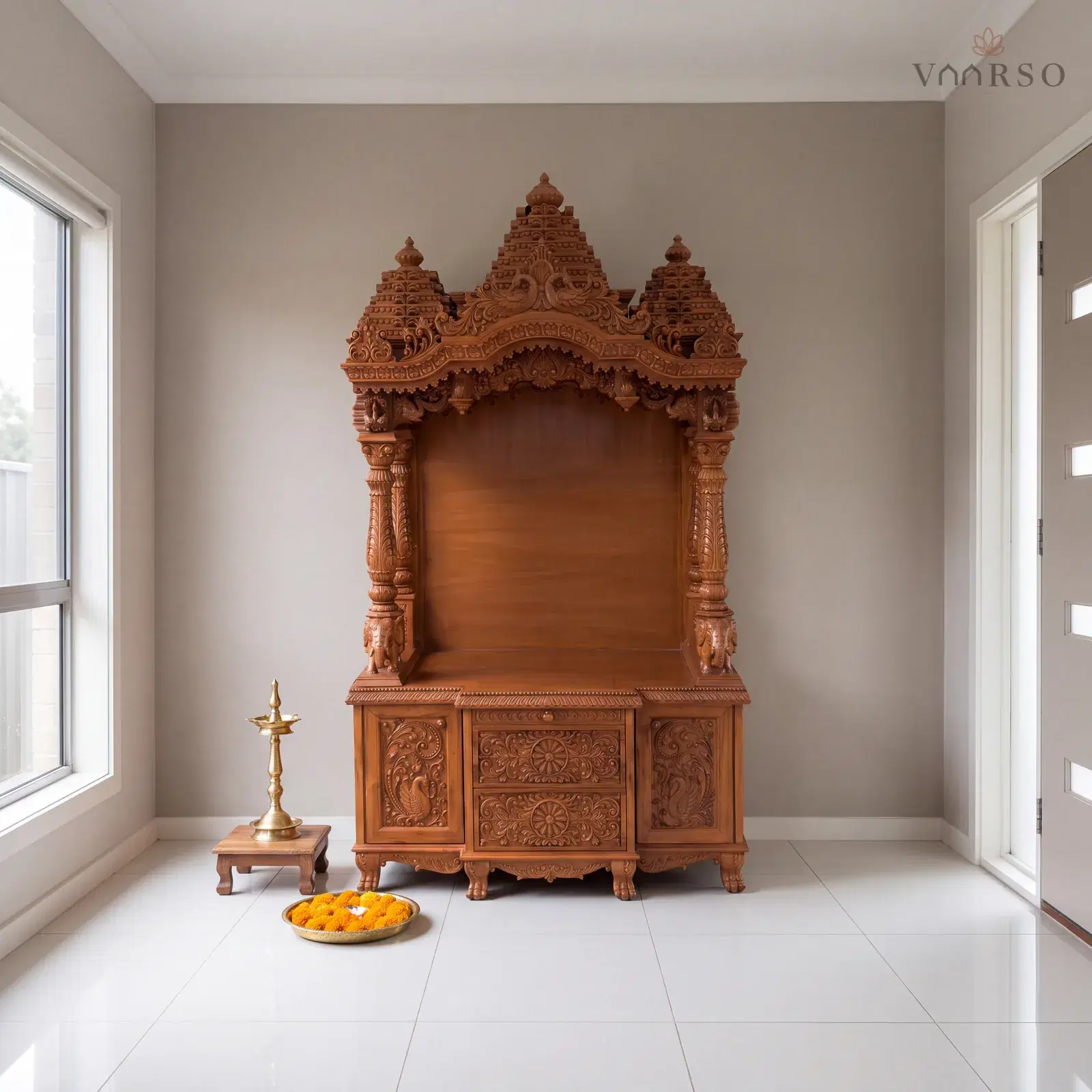The same mandir in an entry foyer, the front door and its sidelight to one side, white porcelain tile underfoot and a brass oil lamp on a timber stool and a thali of marigolds on the floor