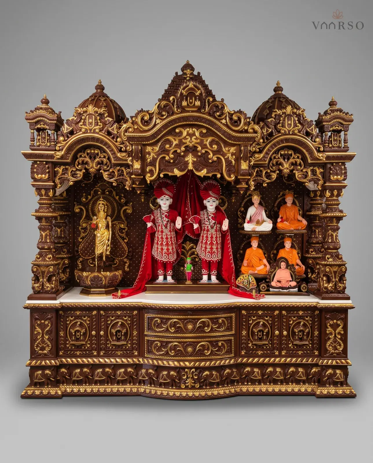 A wide mandir in a dark brown polish picked out in gold, domes and kiosks across the crest, murtis in red dress at the centre with a gilt figure and seated figures in the side bays, and carved drawers over a band of elephants