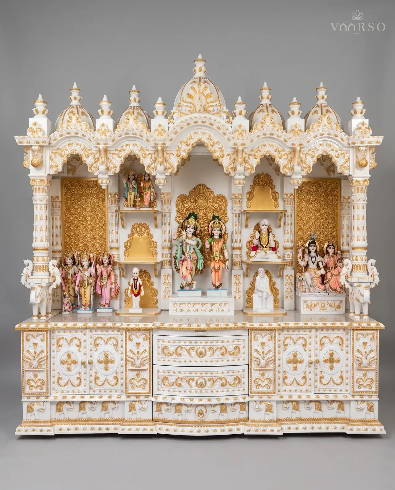 A wide mandir in white polish picked out in gold, five domes over three arched bays, murtis standing across gold-patterned backboards with elephants at the outer columns, and drawers between gilded cupboard doors