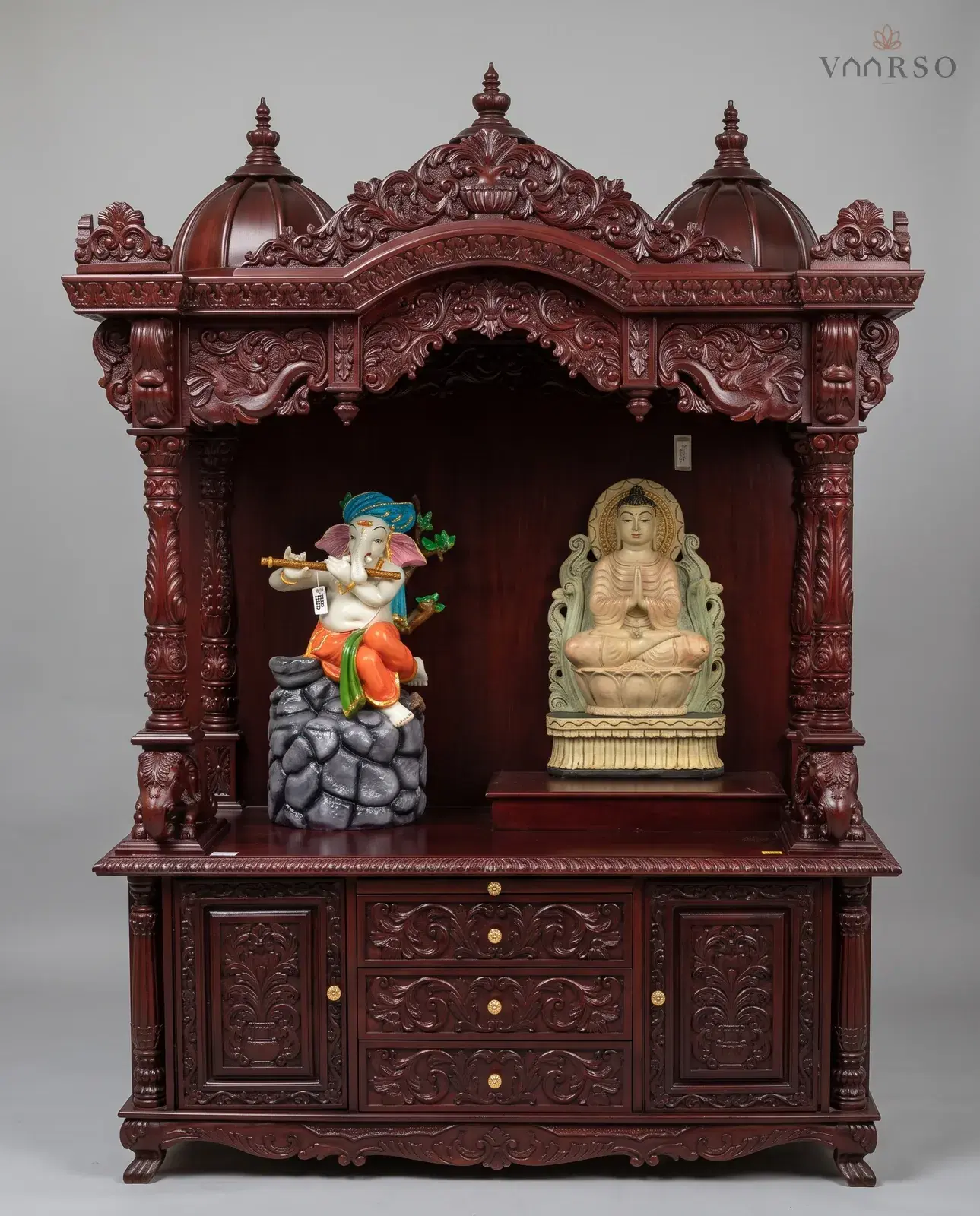 A wide mandir in a deep red-brown polish, three domes over a scrolled valance, a Ganesha and a seated Buddha standing in the open shrine, elephants at the columns, and three drawers between two carved cupboards