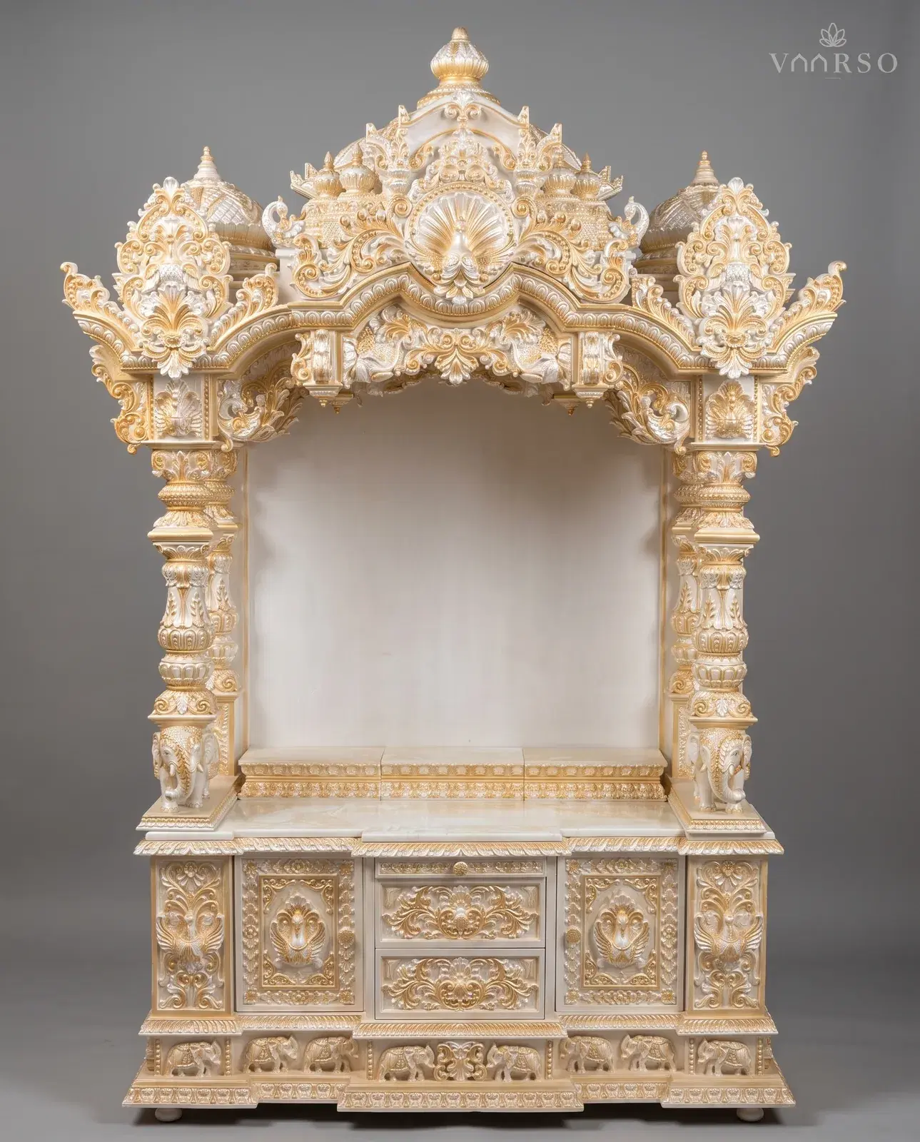 A wide mandir in pearl-white polish picked out in gold, a peacock at the centre of the crest between two domes, an empty shrine over a marble shelf, elephants at the columns, and drawers between carved cupboards over a band of elephants