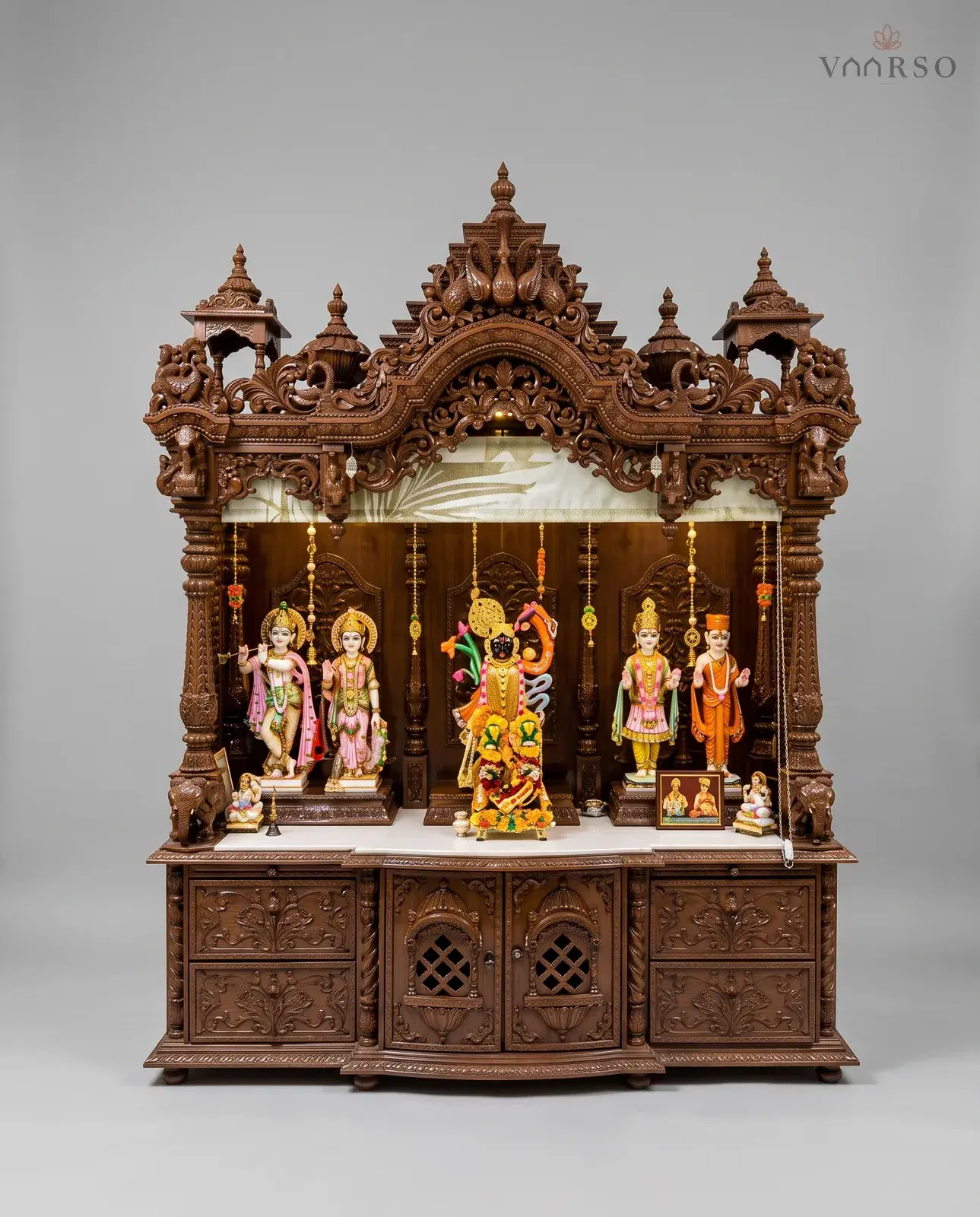 A wide mandir in a dark walnut polish, a stepped crest with peacocks between two kiosks, a printed valance and hanging bead strings over the shrine, murtis standing along a white marble shelf, and drawers beside two latticed cupboard doors