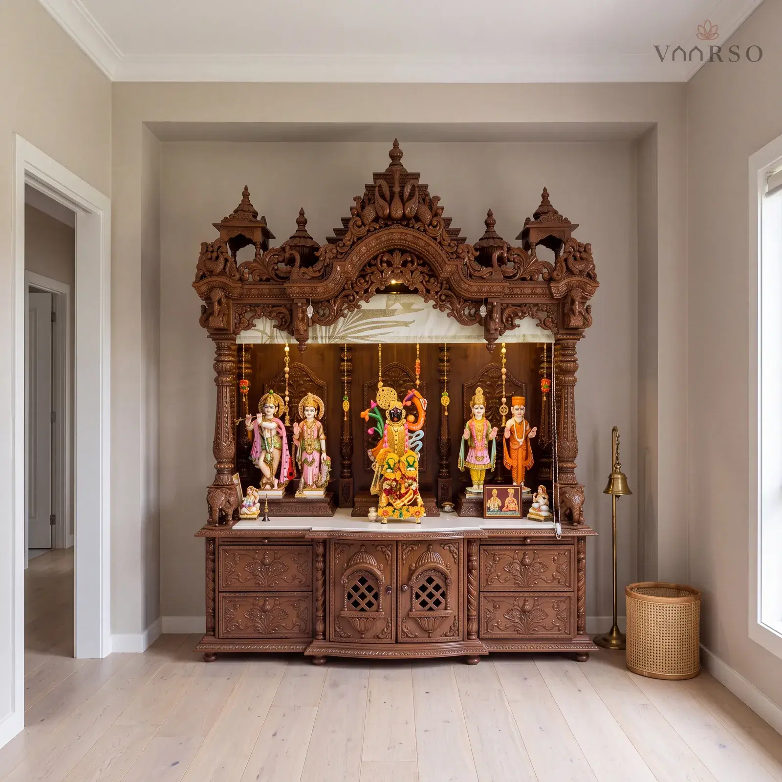 The same mandir in a small pooja room, standing in its alcove, wide pale oak boards underfoot and a brass bell on a floor stand and a cane basket beside it