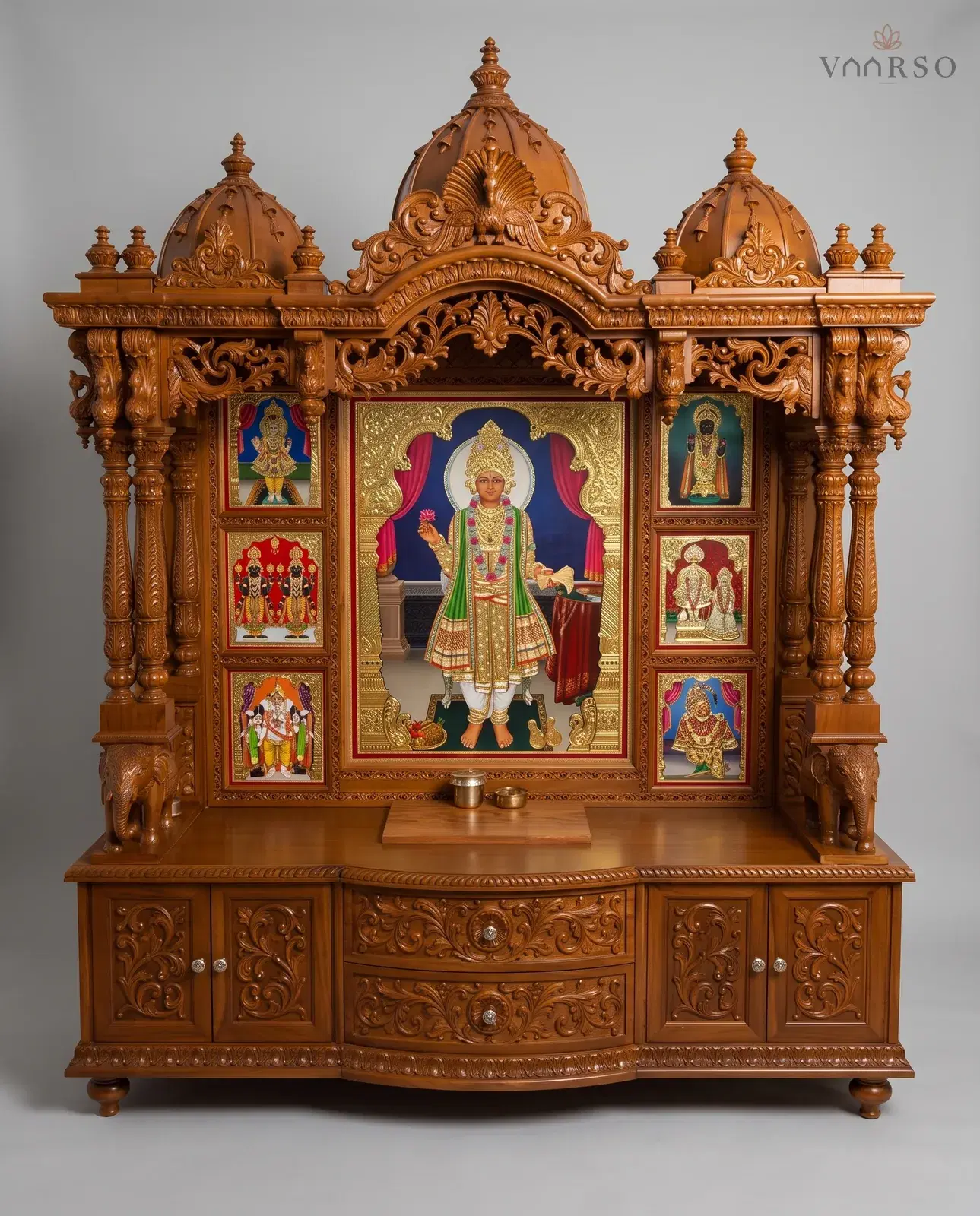 A wide mandir in a warm natural polish, three domes over a pierced valance, a large gilded picture at the centre framed by six smaller ones, elephants on the side brackets, and drawers between carved cupboard doors