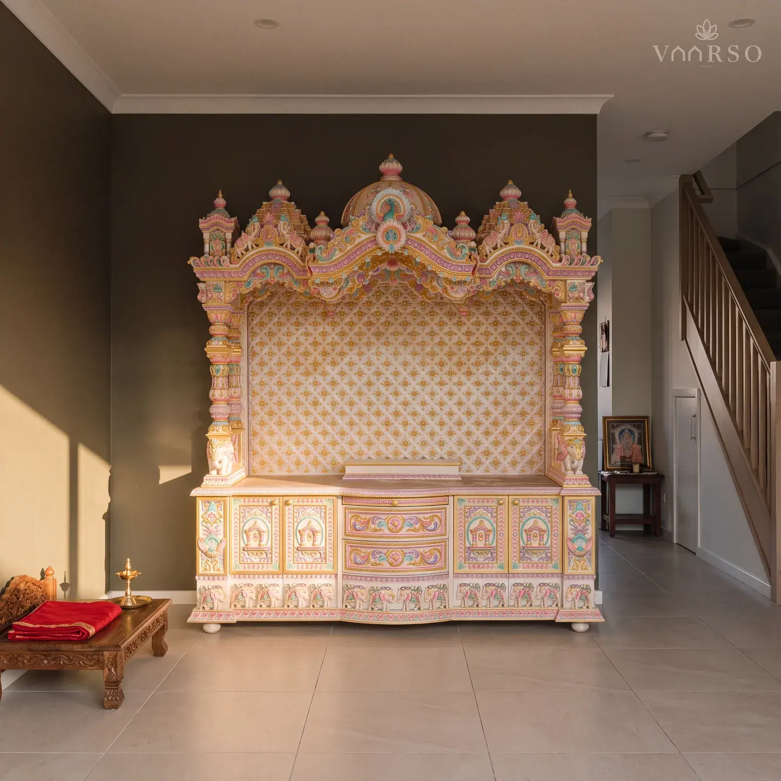 The same mandir in a narrow townhouse living room, the stair rising at the far end, sand-toned porcelain tile underfoot and a low carved chowki with a folded red cloth on it