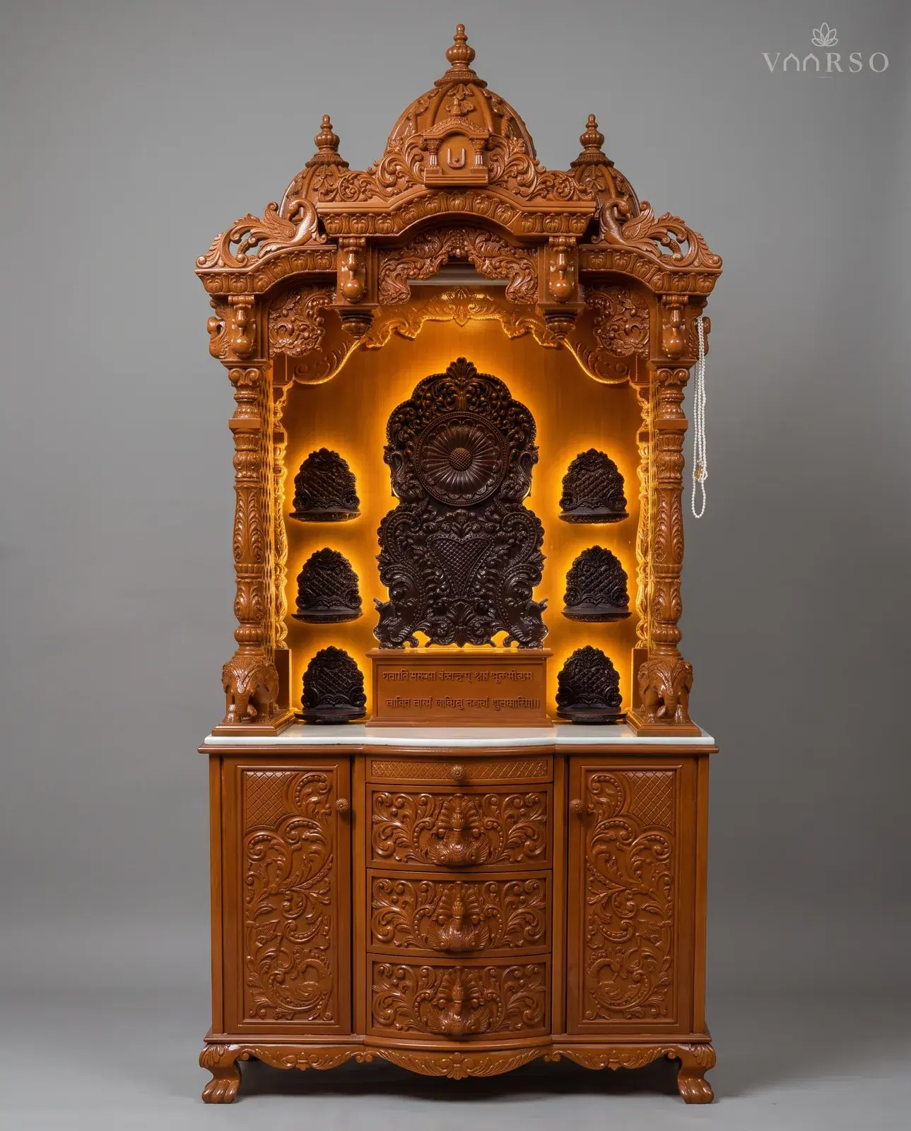 A tall mandir in a warm orange-brown polish, a domed crest over scrolled brackets, a dark carved backpiece lit from behind between six small niches, a white marble shelf, and three drawers between carved cupboards on claw feet