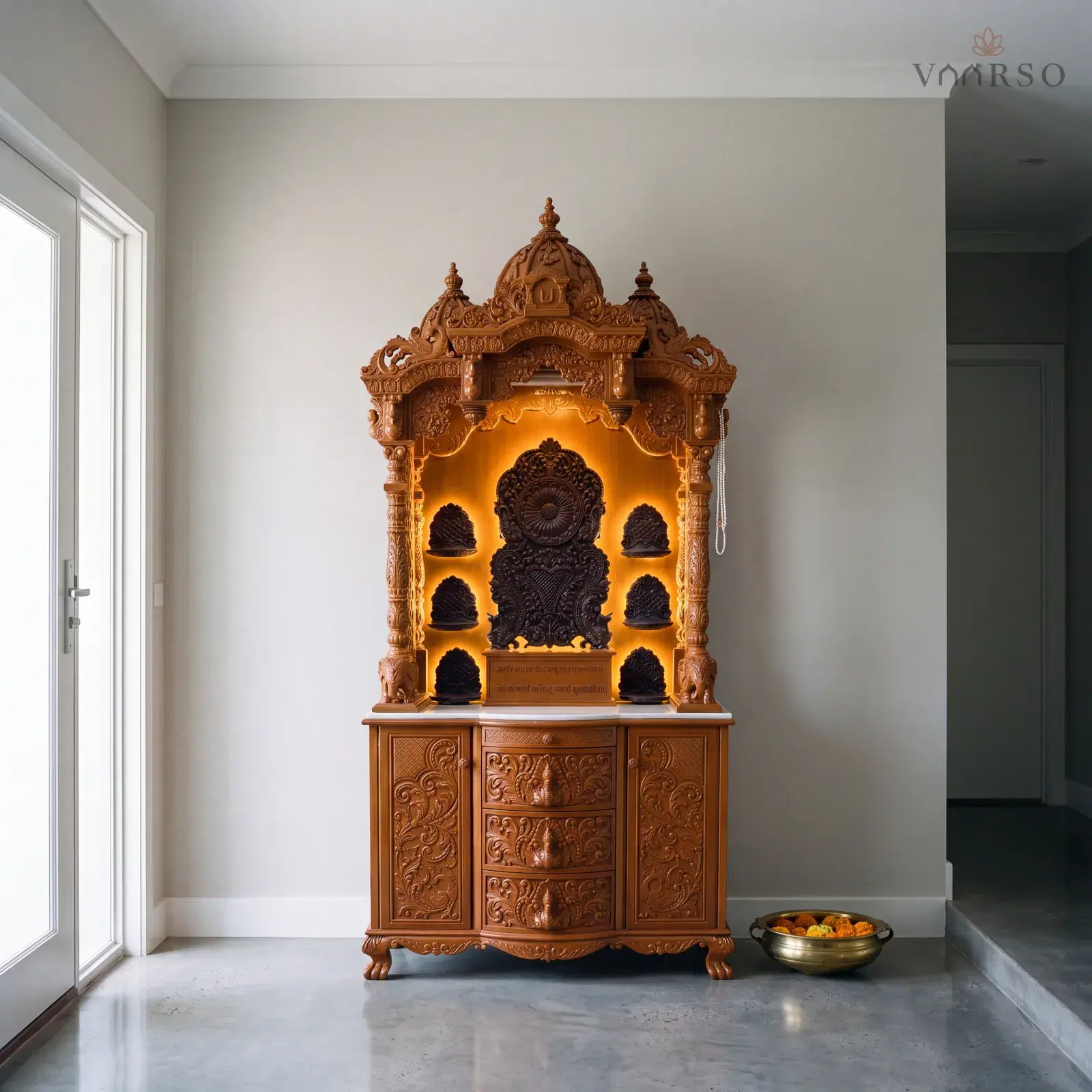 The same mandir in an entry foyer, the front door and its sidelight to one side, polished concrete underfoot and a brass urli of floating marigolds on the floor