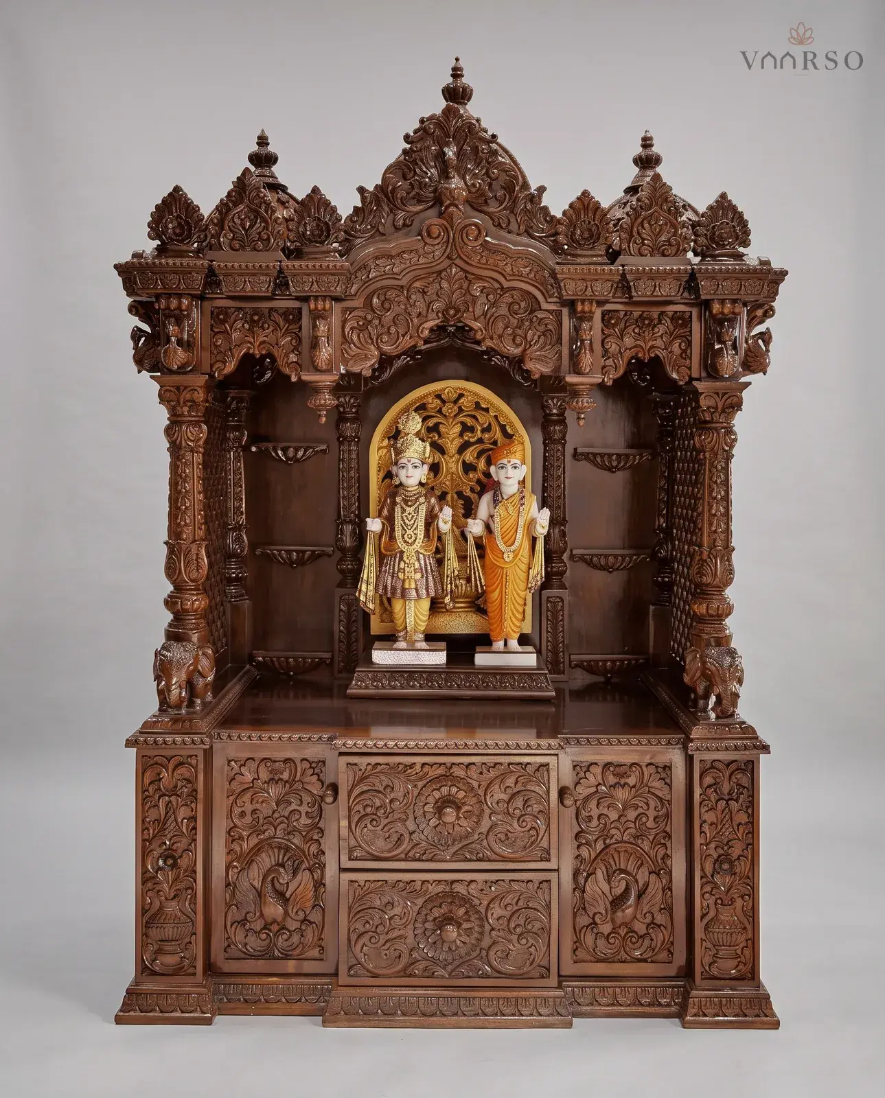 A wide mandir in a dark walnut polish, three crests over a carved valance, murtis standing against a lit pierced panel with empty shelves either side, elephants at the columns, and two drawers between carved cupboards