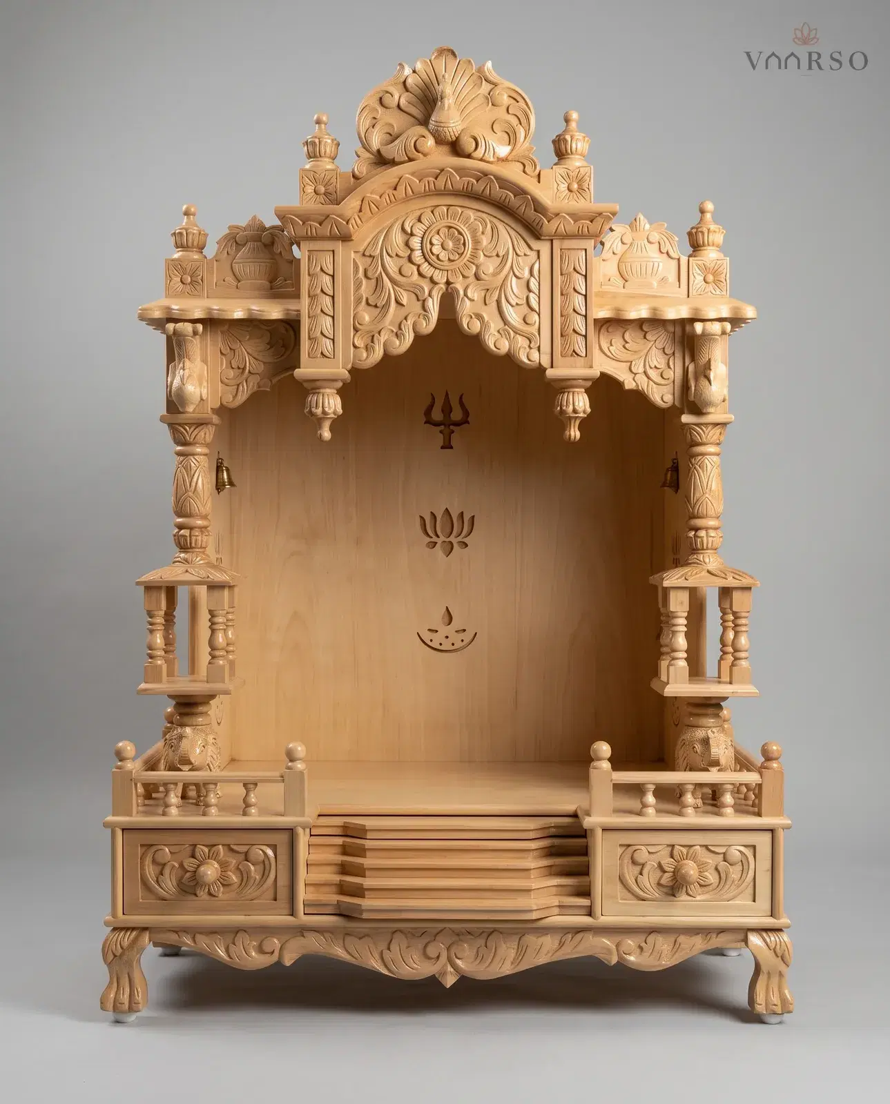 A mandir in pale natural teak, a carved peacock-fan crest over a flower arch, turned columns with small railed balconies, elephants at the base of the columns, centre steps between two carved drawers, and a plain back panel cut with a trident, a lotus and a lamp