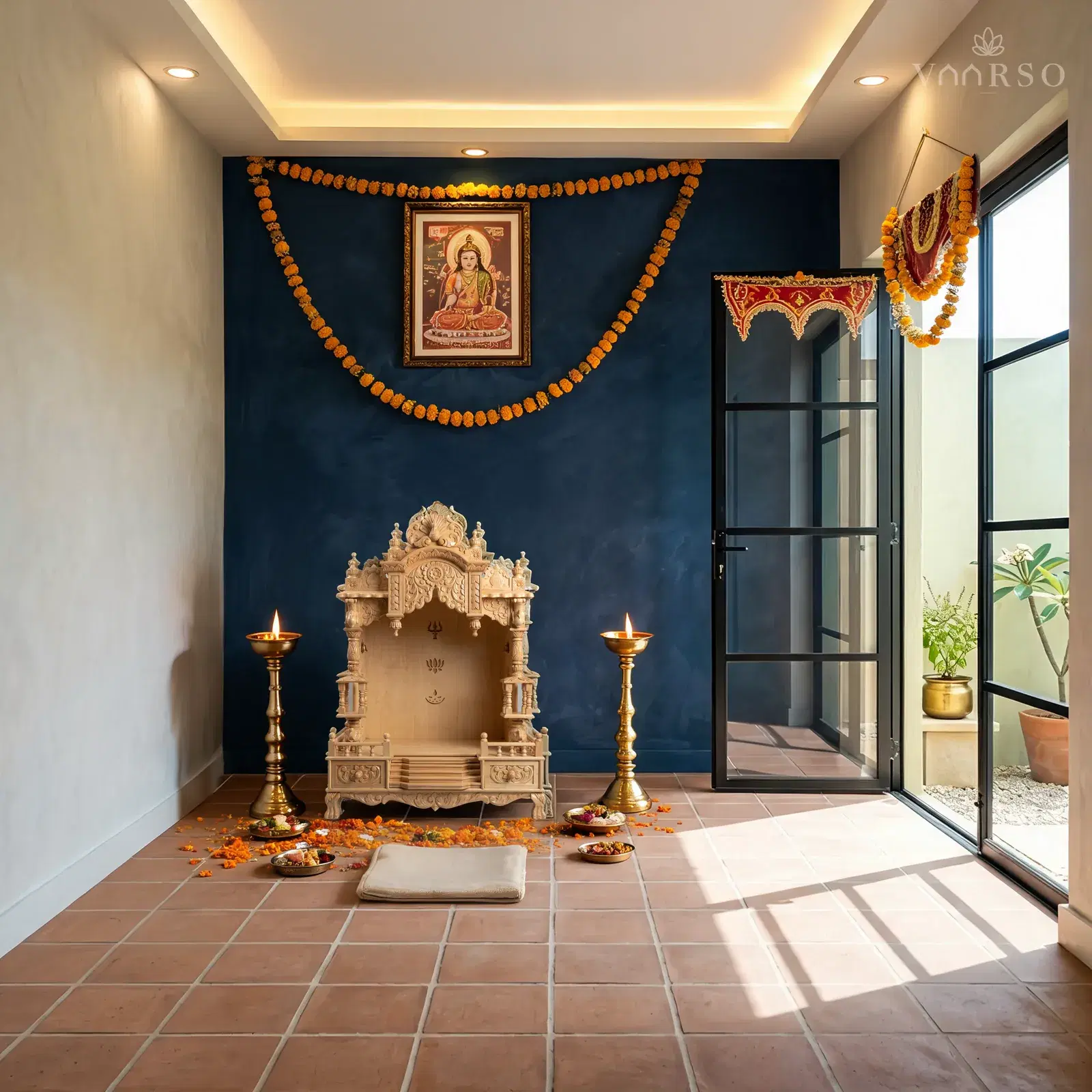 The same mandir against an ink-blue limewashed wall in a courtyard room with terracotta tiles, a lit brass lamp standing on each side, flowers and offerings at its feet, a marigold garland and a framed deity print on the wall above