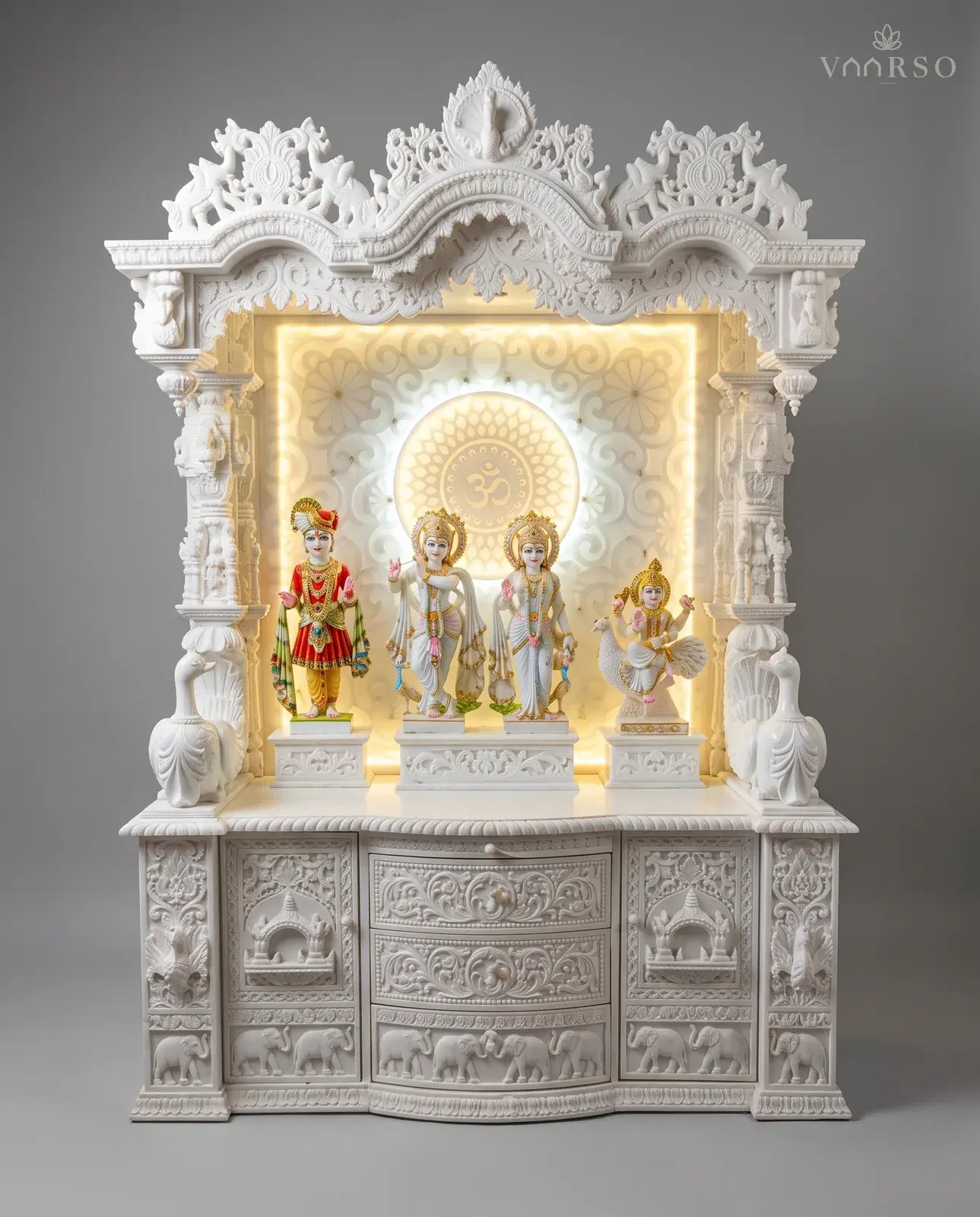 A wide mandir in a white polish, three carved crests over a backlit Om panel, four painted murtis on carved plinths, peacocks at the outer columns, and curved drawers over an elephant frieze