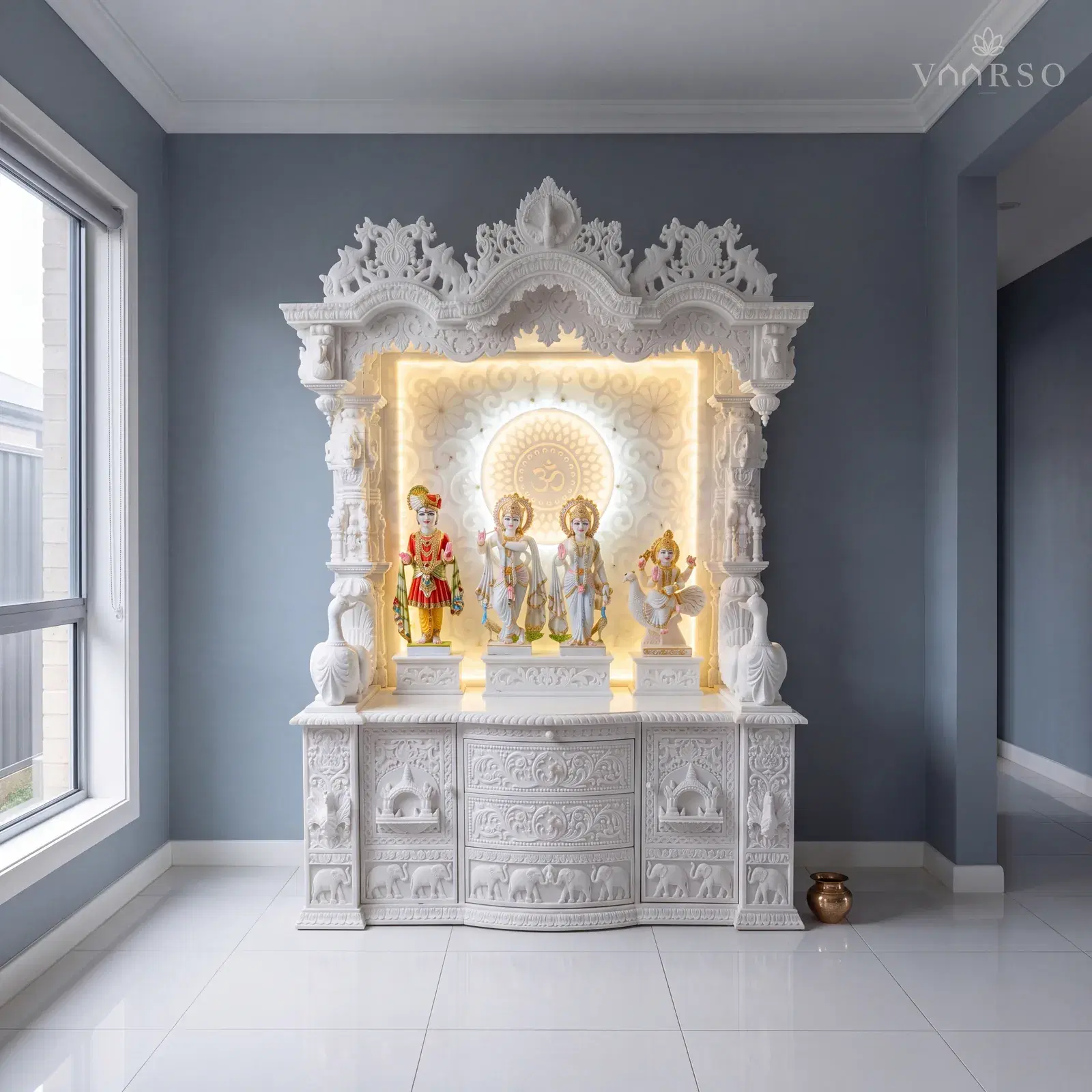 The same mandir in a room with a tall window and a pale tiled floor, a brass pot standing beside it