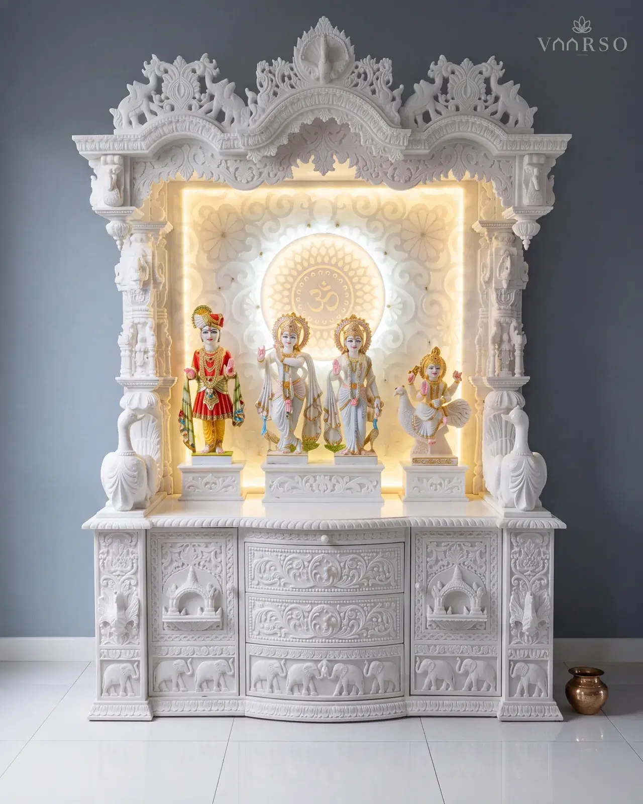 The mandir closer against a grey wall, showing the backlit panel behind the murtis and the carved elephant frieze across its base