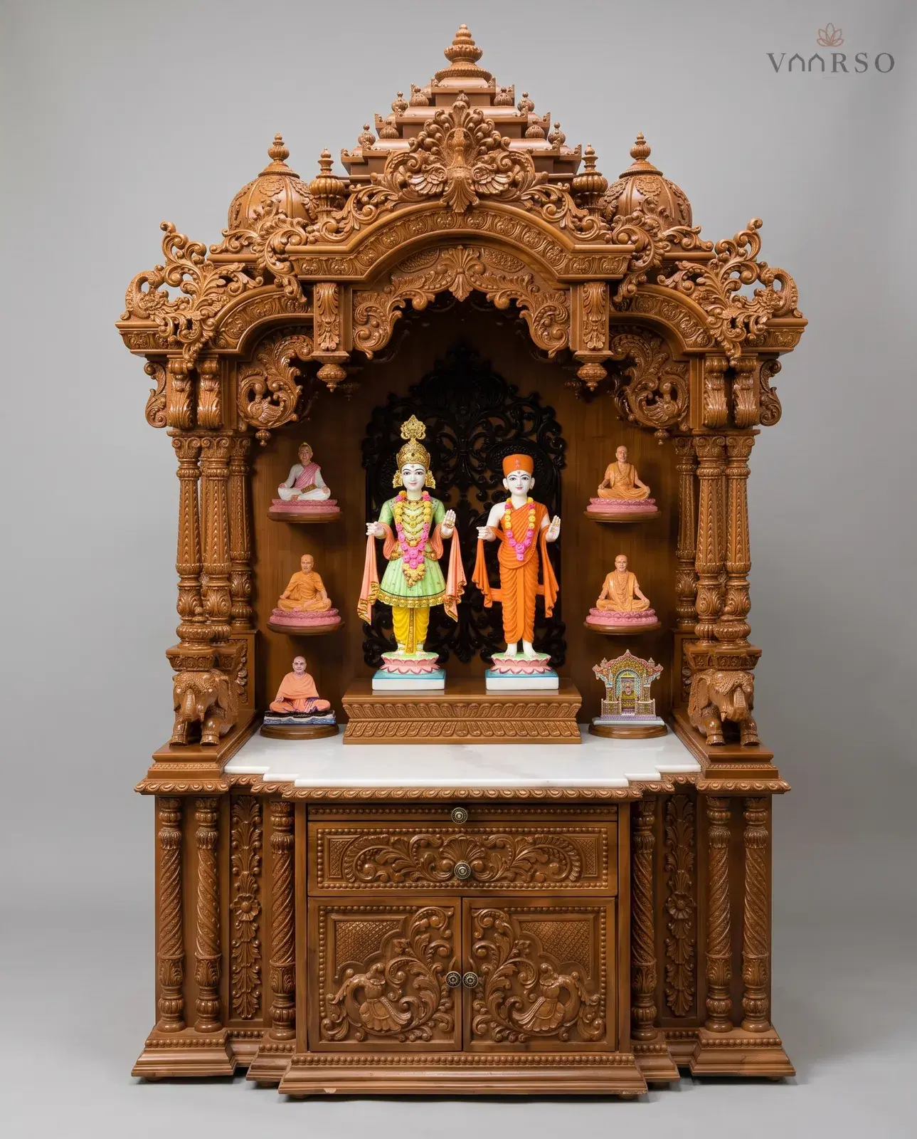 A tall mandir in a warm natural polish, a stepped crest and two domes above a scrolled arch, elephants at the inner columns, a white marble shelf, and a carved drawer over two cupboard doors with brass knobs