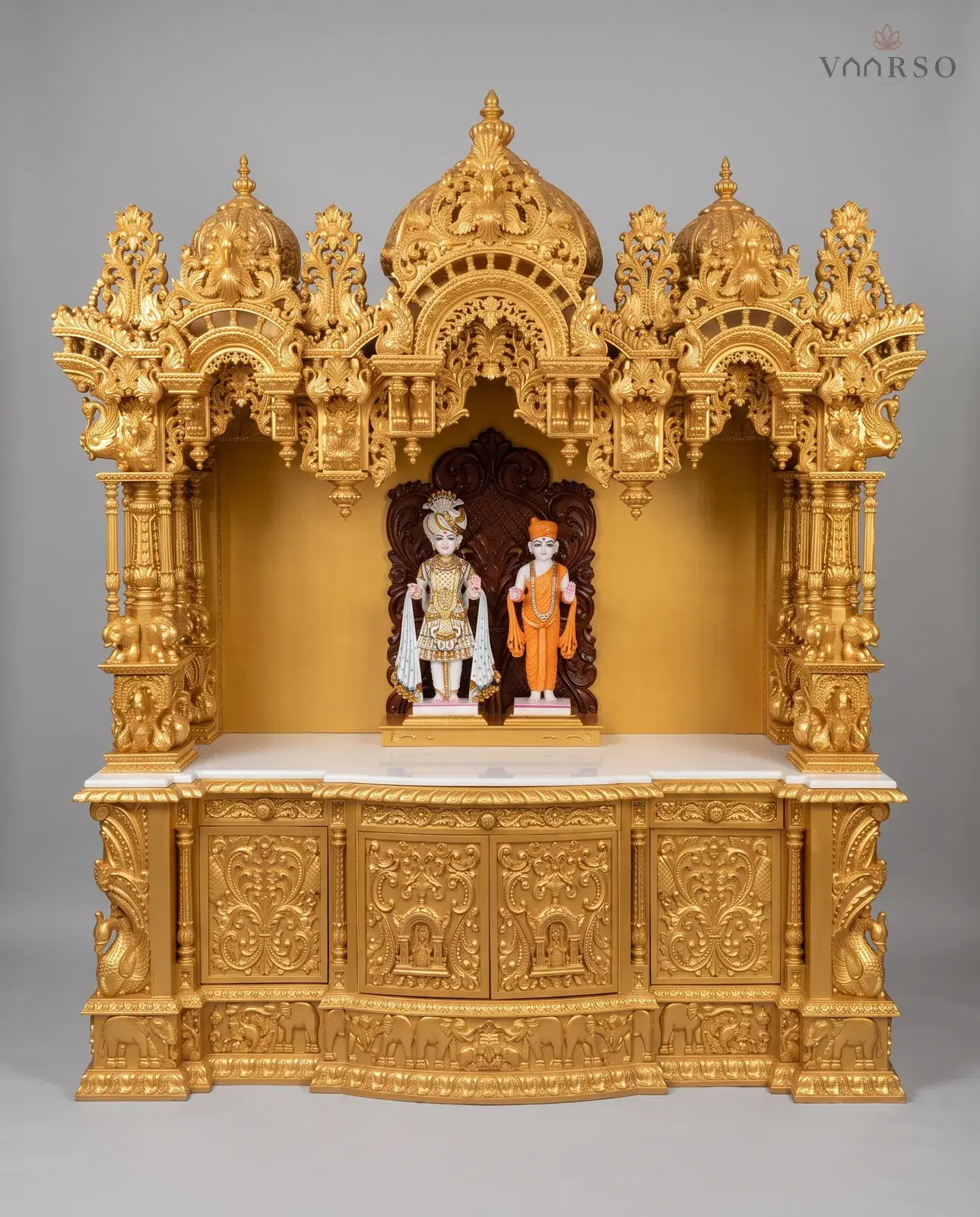 A wide mandir finished in gold, three domes over pierced crests, murtis standing against a dark carved back panel on a white marble shelf, gilded cupboard doors below and a band of elephants around the plinth