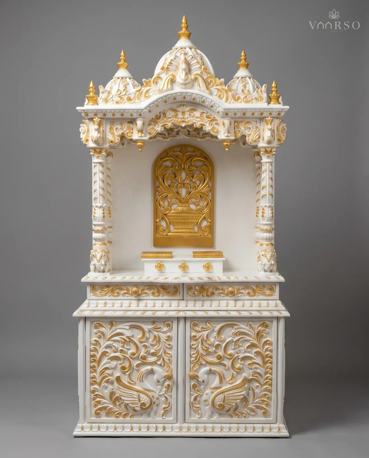 A mandir in white polish picked out in gold, three domes with gilded finials, a gold pierced panel at the back of the shrine, elephant heads on the columns, and two drawers over cupboard doors carved with peacocks