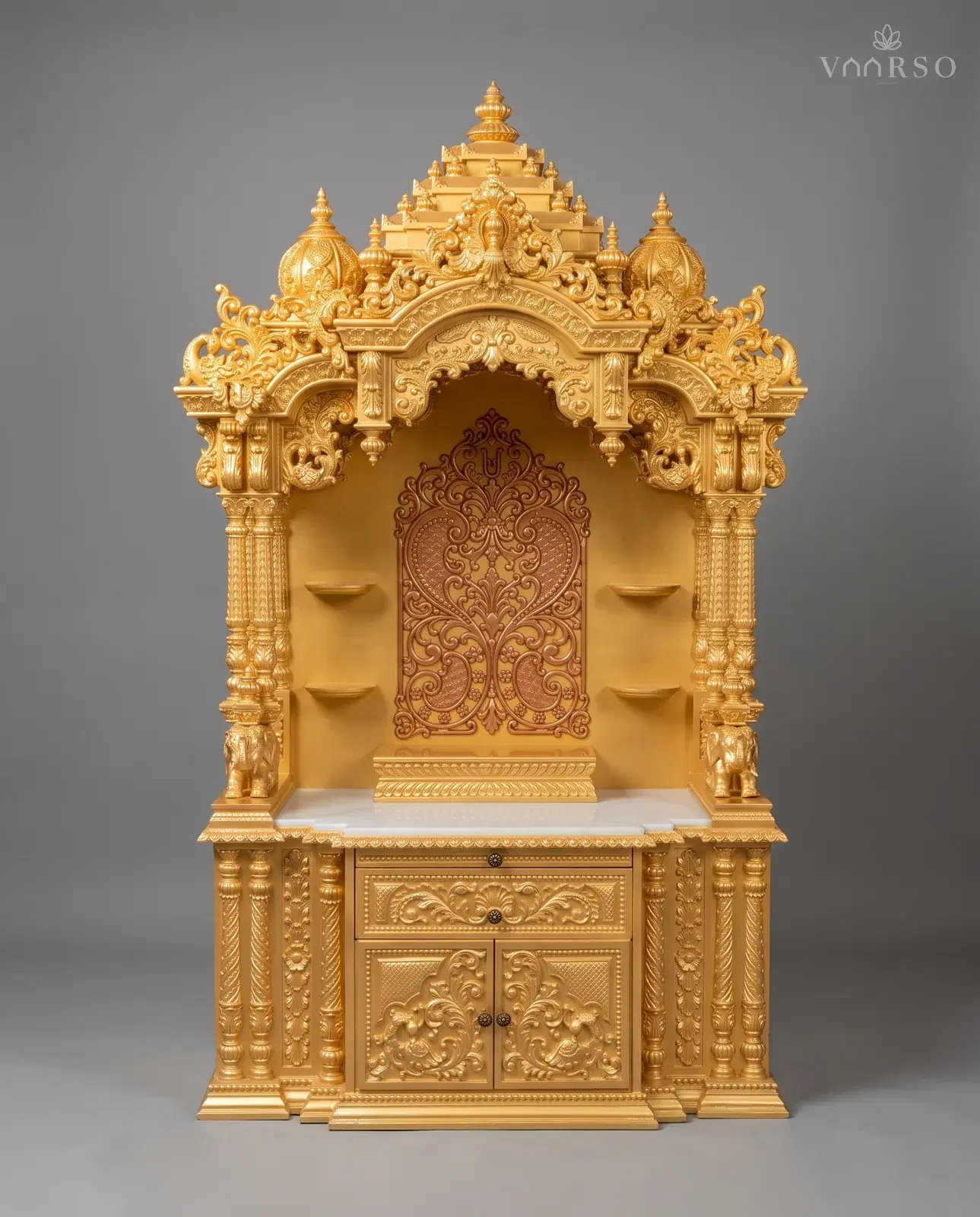 A tall mandir finished in gold, a stepped crest and two domes over a scalloped arch, a copper-toned pierced panel at the back of the empty shrine, a white marble shelf, and a drawer over peacock-carved doors