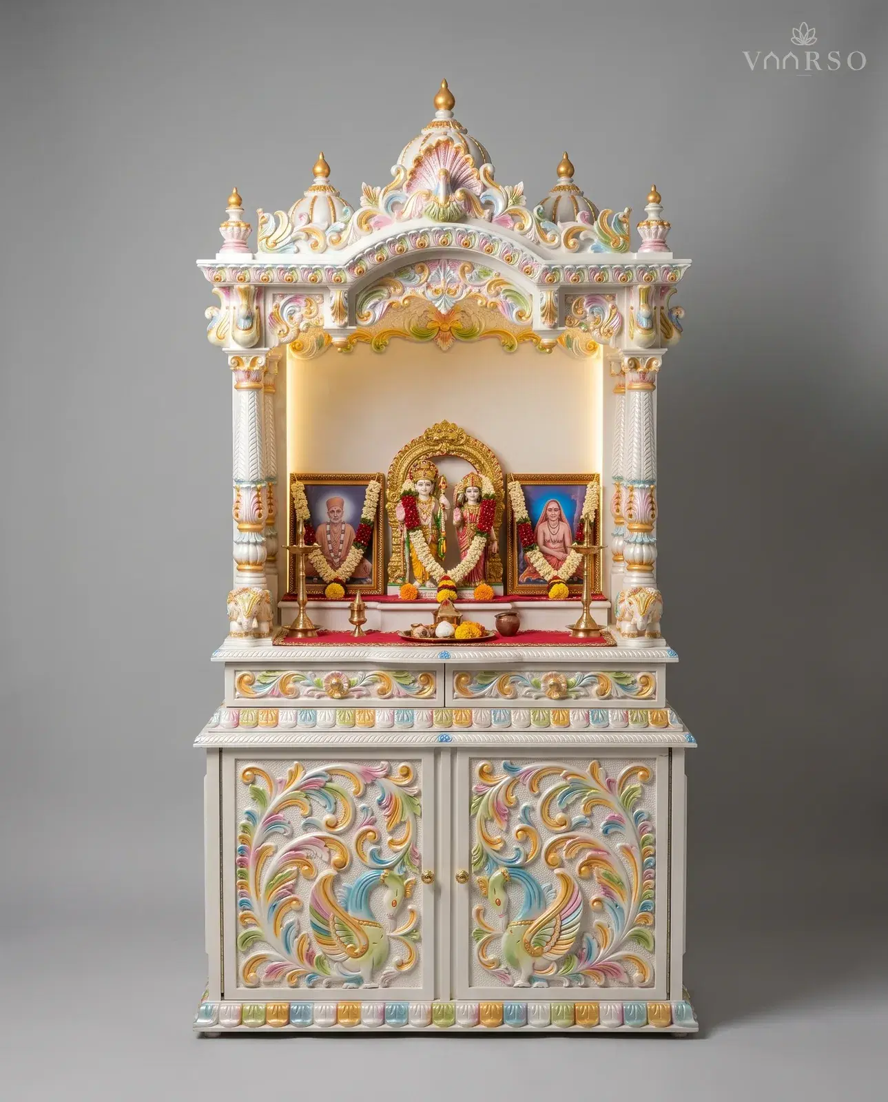 A mandir in white polish picked out in pastel colours and gold, a backlit shrine holding garlanded murtis between two framed portraits, brass lamps on the shelf, and cupboard doors carved with peacocks