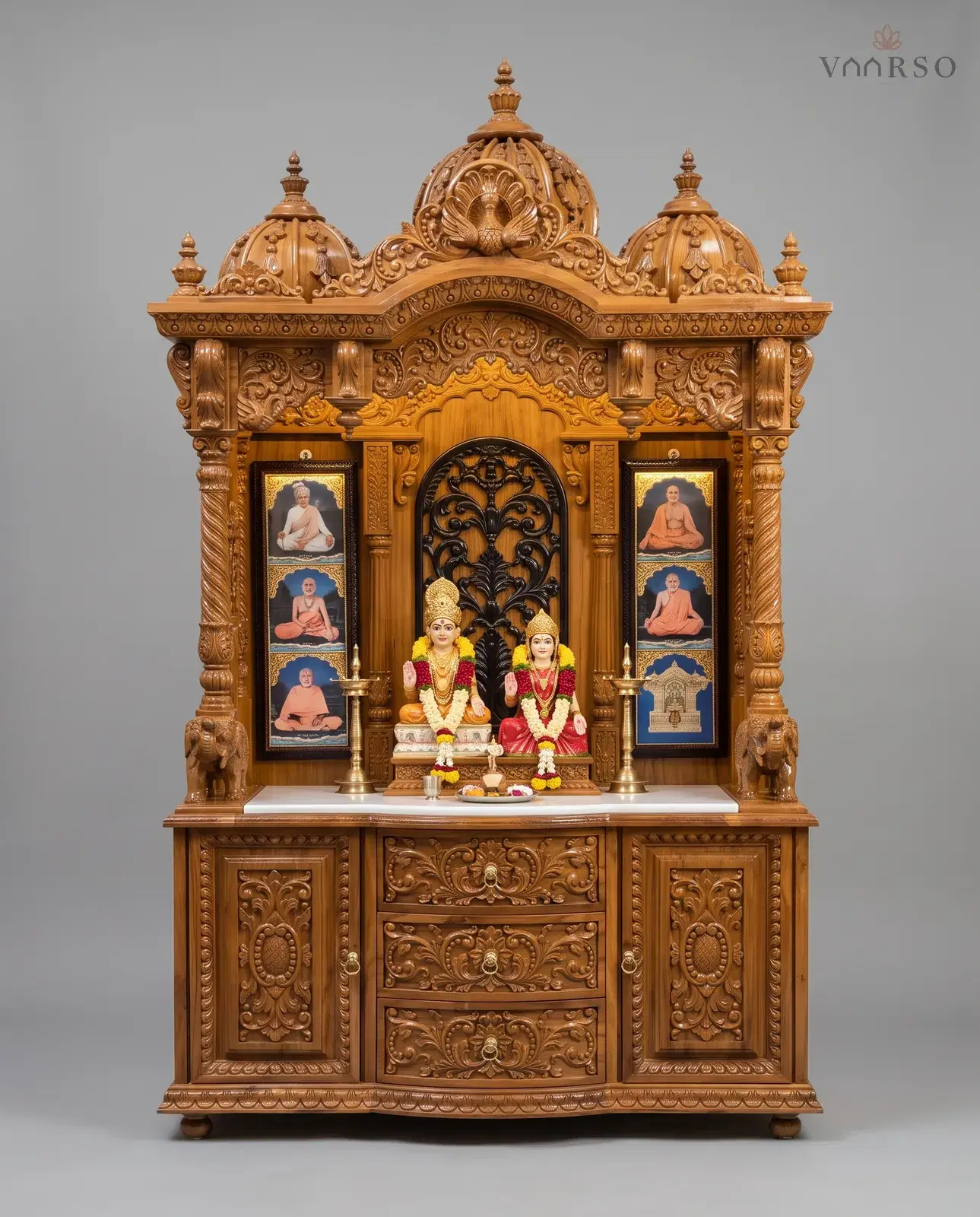 A wide mandir in a warm natural polish, three domes over a carved valance, painted panels either side of a dark pierced backpiece, a white marble shelf, and three drawers between carved cupboards
