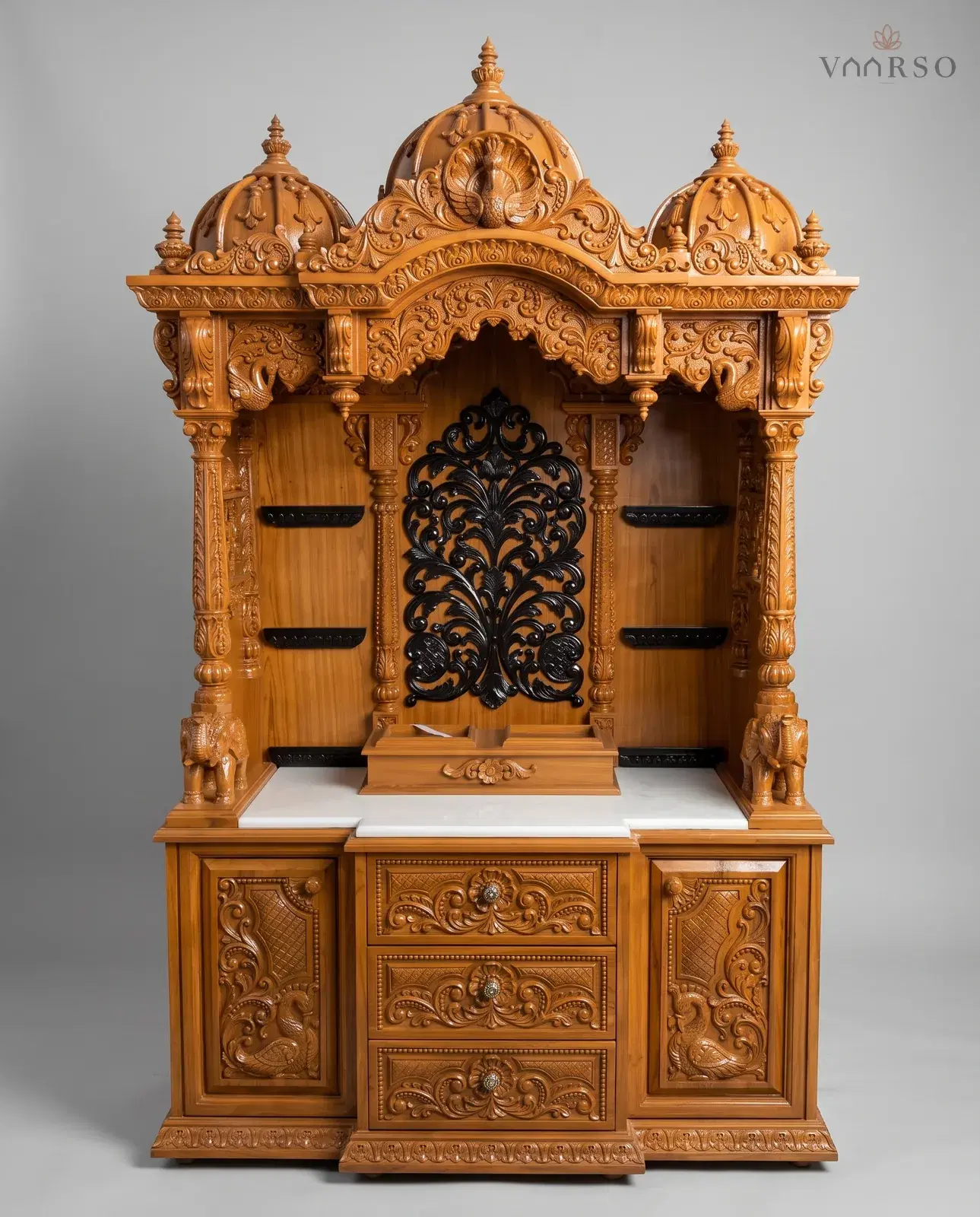 A wide mandir in a warm natural polish, three domes with peacocks over a scalloped arch, a black pierced panel at the back of the shrine between black shelves, elephants on the marble shelf, and three drawers between carved cupboards
