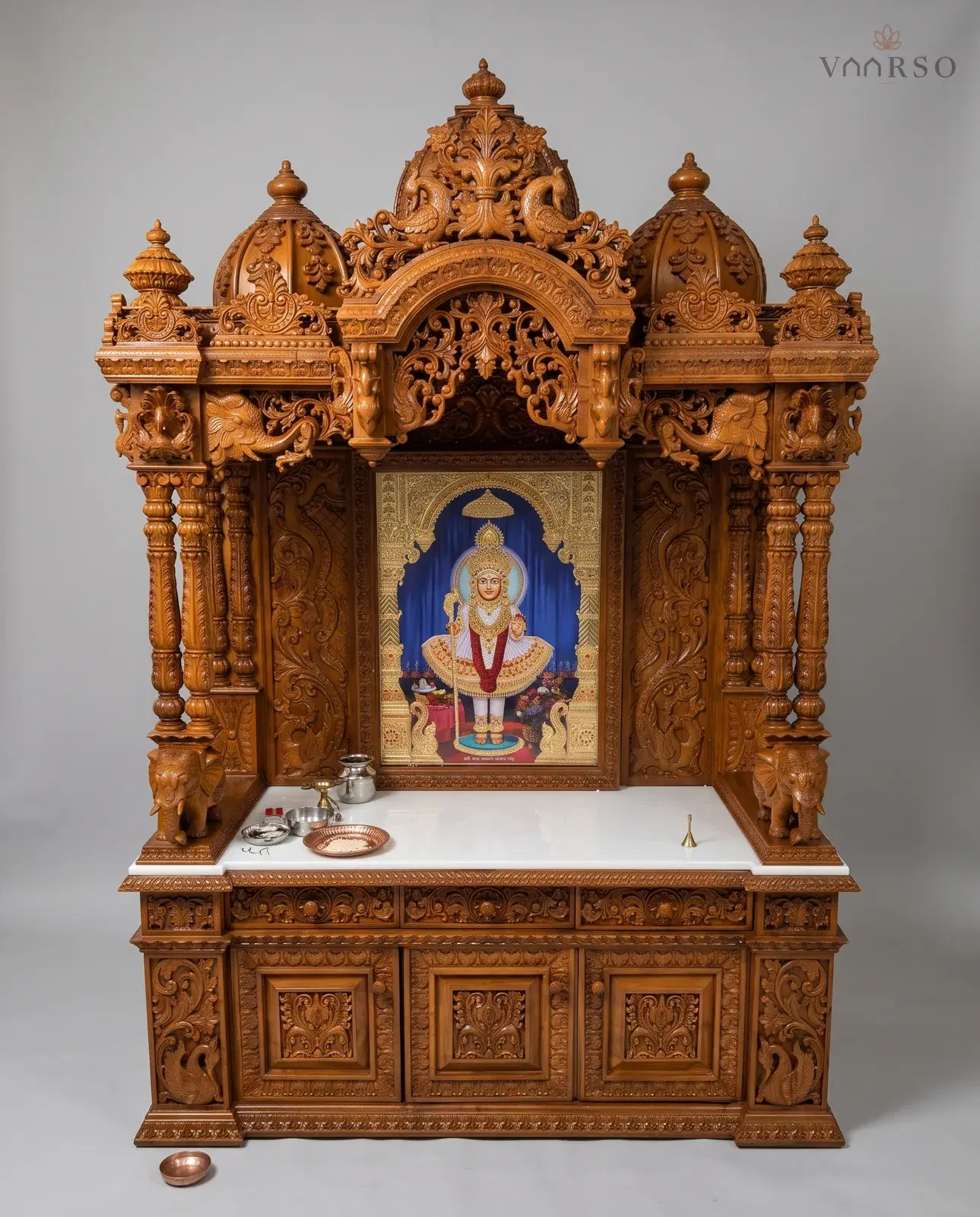 A wide mandir in a warm natural polish, a domed crest over a carved arch, elephants and peacocks at the columns, a framed picture standing on a white marble shelf, and a row of drawers over three panelled cupboard doors
