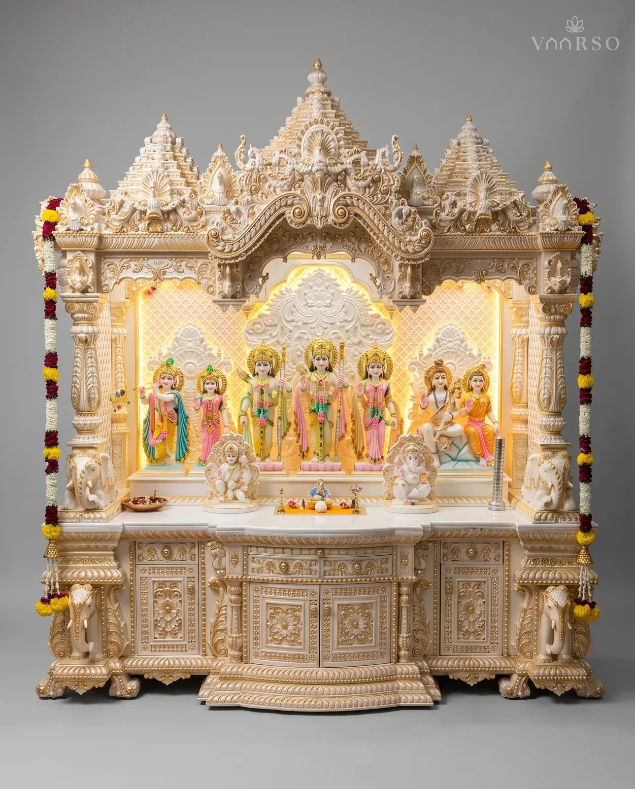 A wide mandir in cream and gold, three stepped shikhars over a scrolled crest, a backlit shrine of many murtis with Ganesha and a seated figure on the shelf, elephants at the columns, and flower garlands hanging down both sides