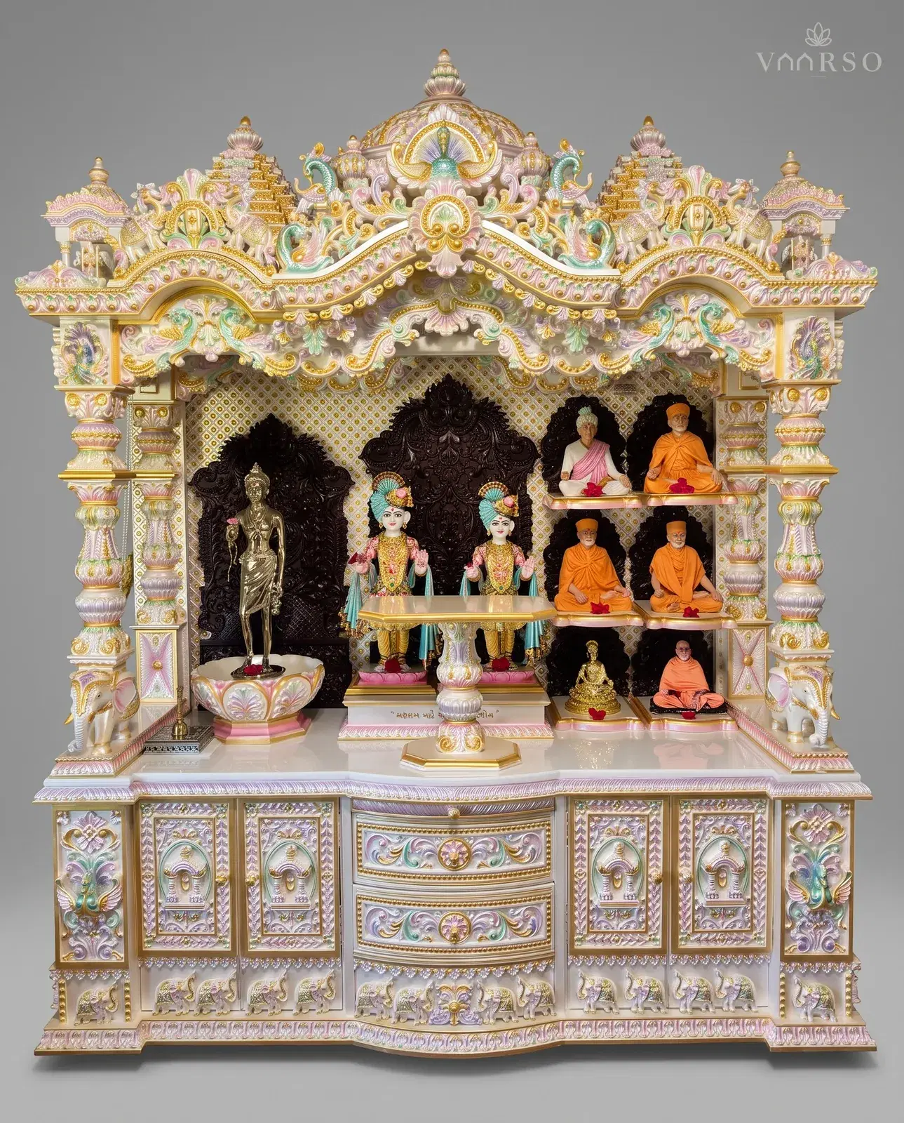 A wide mandir in pearl-toned polish with gold and pastel detailing, shikhars and peacocks across the crest, murtis under a carved canopy with seated figures on the side shelves, and carved drawers over a plinth of elephants